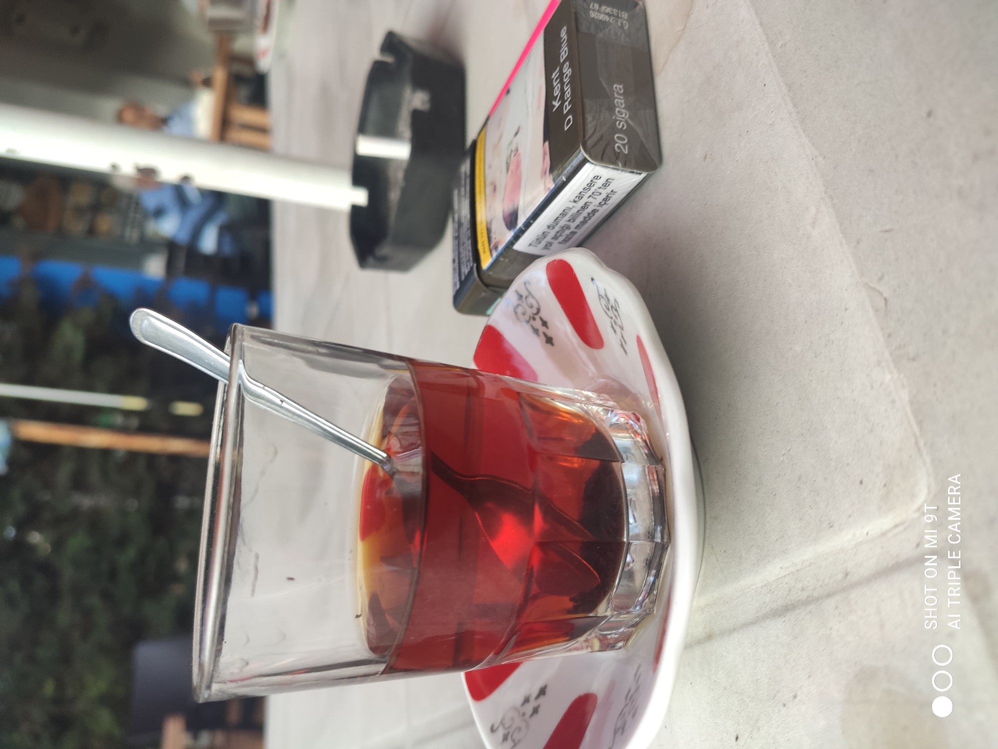 A close-up, eye-level shot shows a glass of dark tea on a saucer with red and white designs. A silver spoon rests in the tea. To the right of the glass is a pack of Kent D Range Blue cigarettes, showing the text "20 sigara." Behind the cigarette pack is a black ashtray. The scene is set on a textured white surface, likely a table. In the blurred background, trees and structures are visible, suggesting an outdoor setting in Akçaabat, Turkey. The bottom left corner of the image displays "SHOT ON MI 9T AI TRIPLE CAMERA."