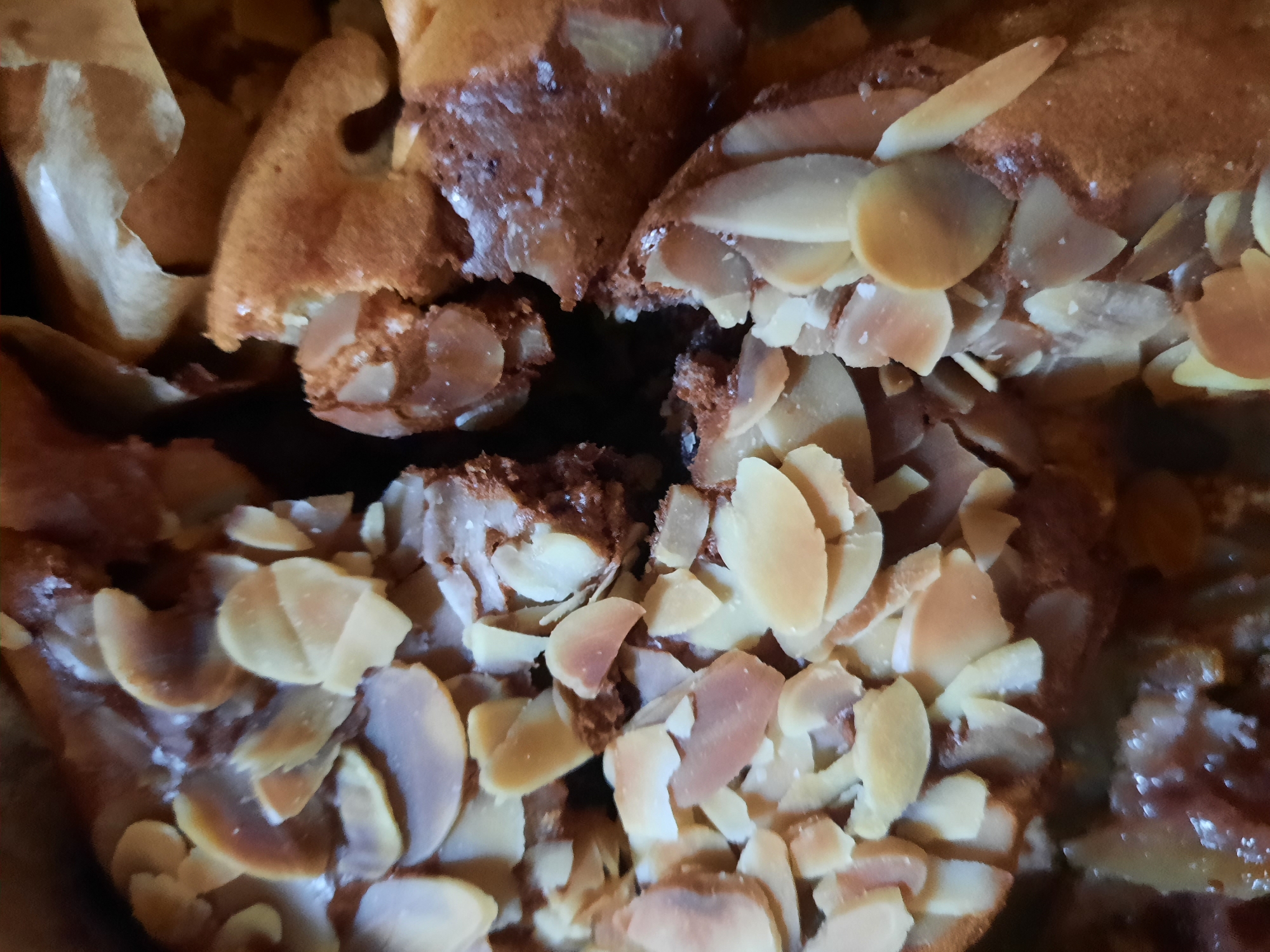The image shows a close-up view of almond pastries, possibly croissants or Danish pastries, with sliced almonds on top. The pastries are golden brown and appear to be freshly baked. The texture of the almonds and the pastry is clearly visible. The background is dark and out of focus, suggesting the pastries are in a dimly lit environment. There are no people or specific landmarks visible to indicate a location within Rosenheim, Germany.