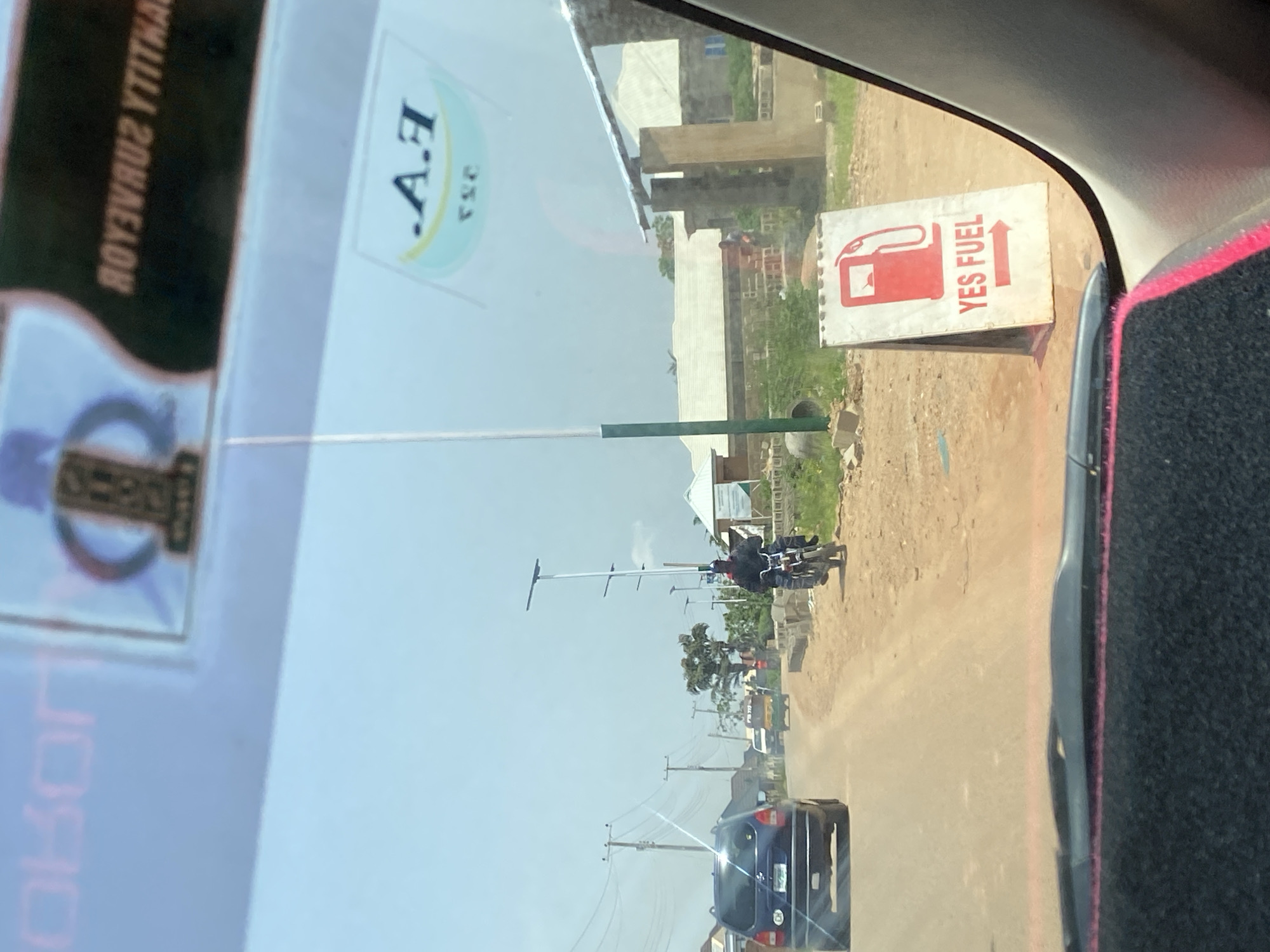 The image, taken from inside a vehicle, shows a street scene in Gura Top, Nigeria, on a clear, sunny day. A blue Opel car is visible on the left, driving on a dirt road. To the right of the Opel, a man on a motorcycle is seen riding towards the viewer. Further down the road, a yellow and green auto-rickshaw is also visible. In the foreground, a white A-frame sign with a red gas pump icon and the text "YES FUEL" with an arrow pointing right is positioned on the side of the road. The background shows houses and vegetation, with several tall light poles lining the street. A sticker with the text ".A.F" and "YSE" is visible on the vehicle's windshield. Another sticker, partially obscured, has text that appears to be in reverse.