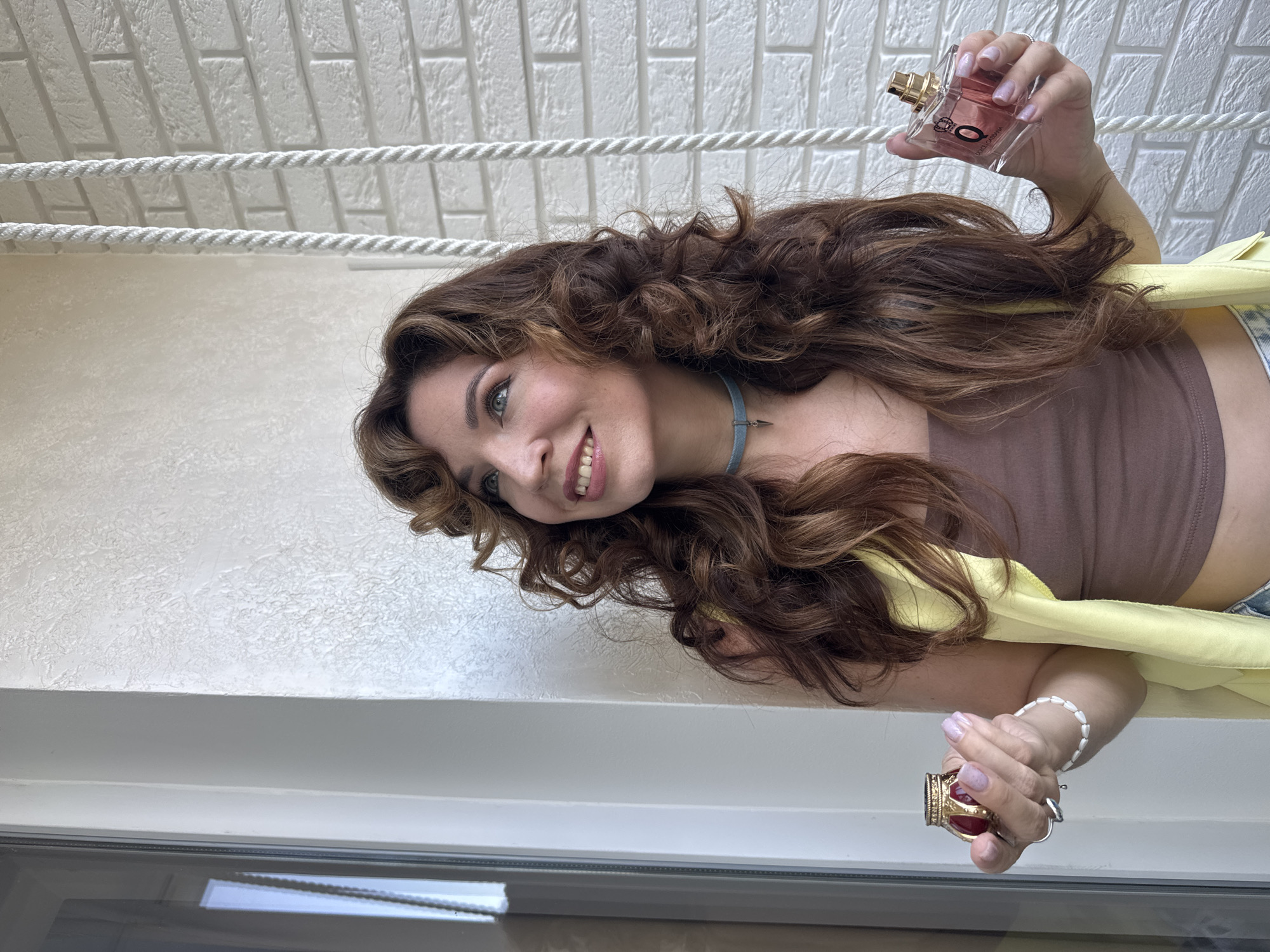 A woman with long, curly brown hair is standing outdoors, holding a small, ornate red and gold perfume bottle in her left hand and a larger, rectangular glass perfume bottle with a pink liquid and a gold cap in her right hand. She is wearing a brown tube top, a yellow vest, and denim shorts. She has a blue choker necklace with a small silver charm. The background consists of a white brick wall with textured plaster to the left, and a window with a grey frame on the far left. Two white ropes hang vertically on the right side of the image. The woman is smiling and looking up and to her left. The lighting suggests it is daytime. The location is not specified, therefore no assumption on the city of Kryvyi Rih, Ukraine, or any other city can be made.