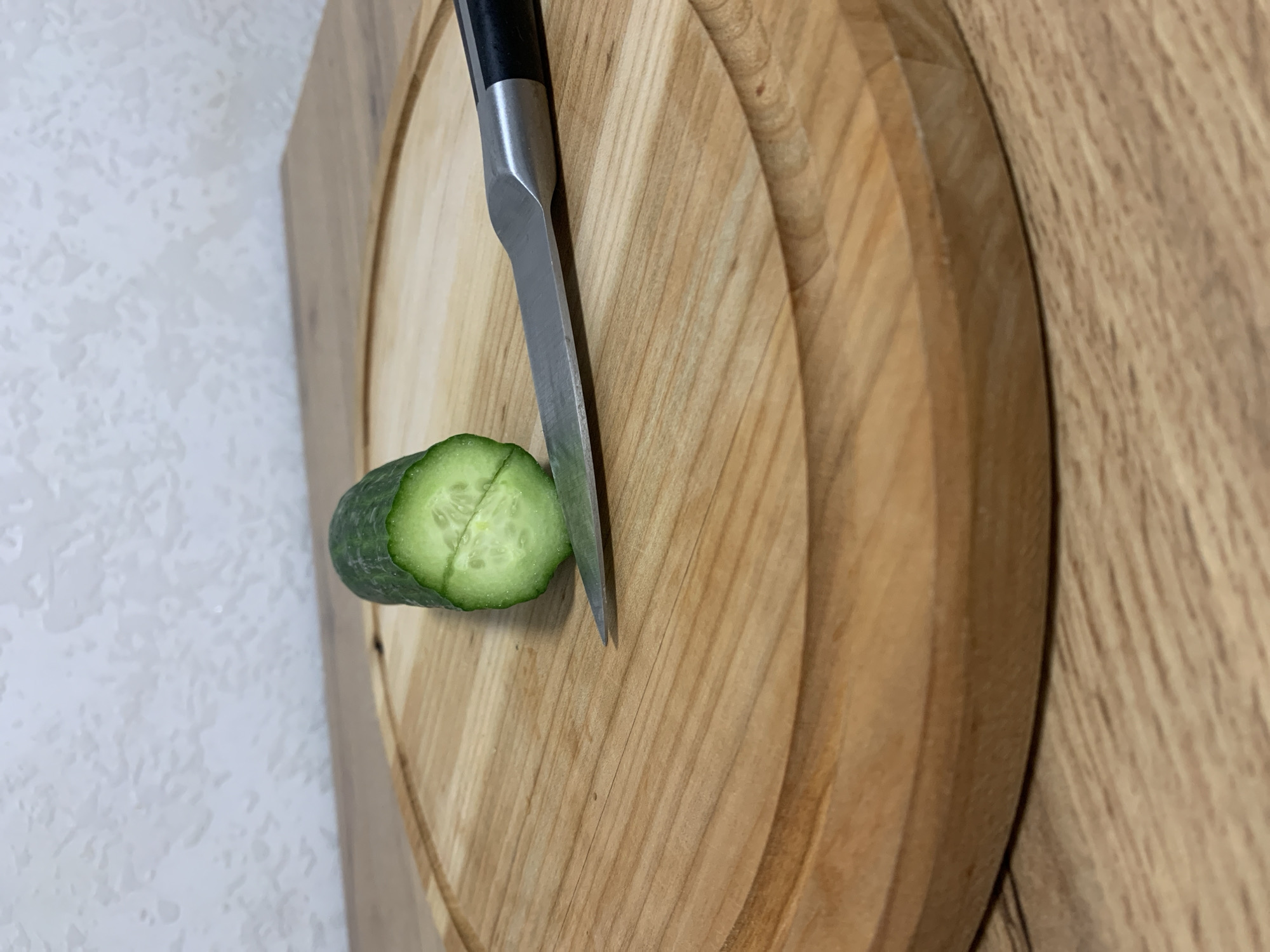 The image captures a close-up scene of food preparation on a wooden surface. The primary subjects are a half-cut cucumber, a kitchen knife, and a circular wooden cutting board. The cucumber is vibrant green, showing its white flesh and seeds where it has been sliced in half. A silver-bladed kitchen knife with a black handle is positioned vertically into the circular cutting board, its tip resting near the cut surface of the cucumber.
The circular cutting board, made of light-colored wood with visible grain, features a shallow groove running along its perimeter. This board is placed on a larger, lighter-toned wooden surface, which could be another cutting board or a countertop. The background consists of a textured, light-colored wall, possibly white or off-white. The lighting appears even, suggesting an indoor setting with artificial illumination. No people, text, or specific time of day are visible. The scene conveys a moment of culinary activity, likely in a kitchen in Ternopil, Ukraine, focusing on the simple act of preparing fresh produce.