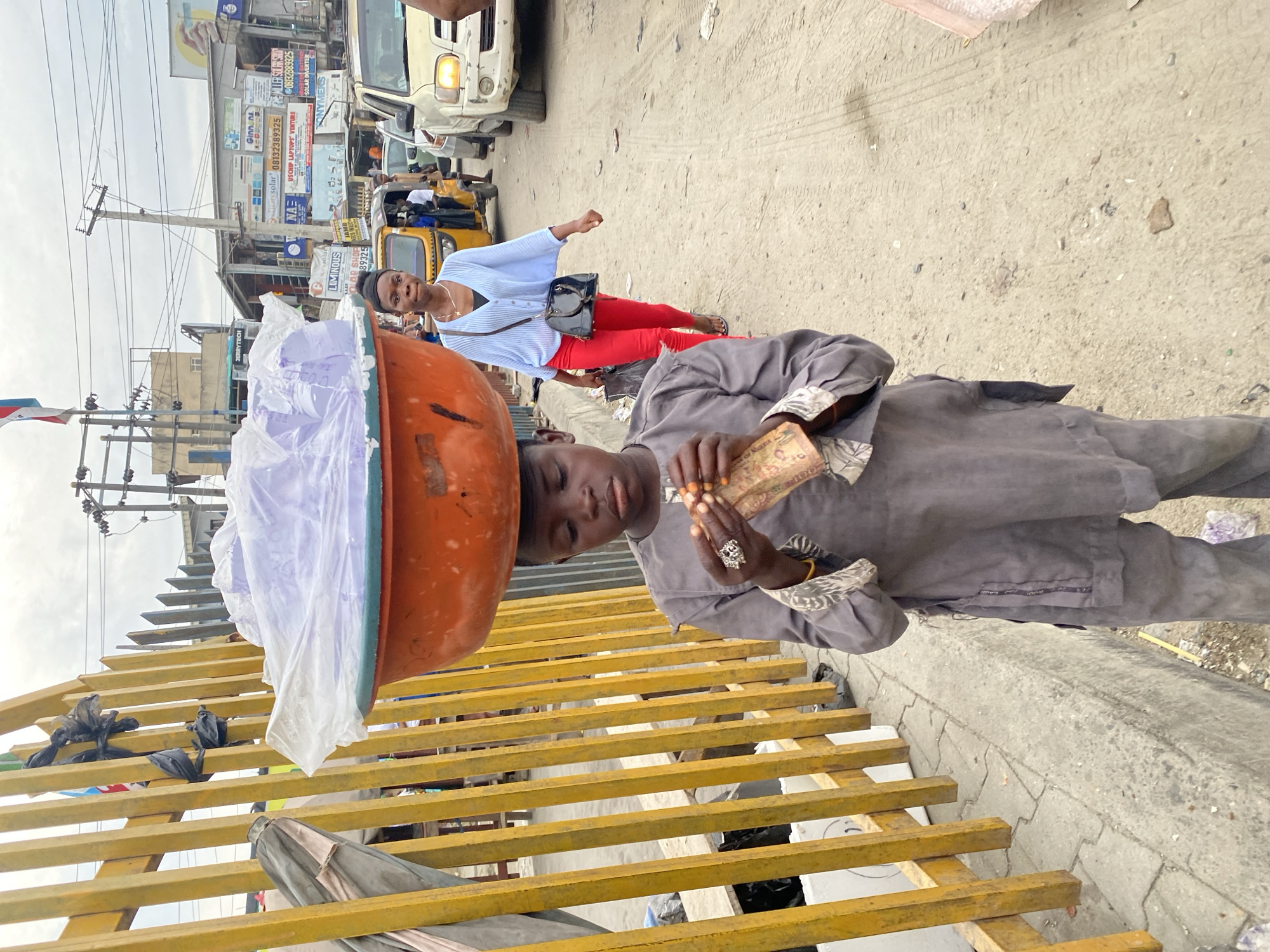 The image depicts a street scene in Lagos, Nigeria. A young person is balancing an orange tray filled with what appears to be bagged water or drinks on their head. They are wearing a grey outfit and holding currency, possibly transacting business. In the background, a woman in a light blue shirt and red pants is walking along the sidewalk. Traffic and various commercial signs are visible, including those advertising solar products and laptops. A tricycle is parked nearby. The area appears to be a busy commercial district with visible power lines and infrastructure.