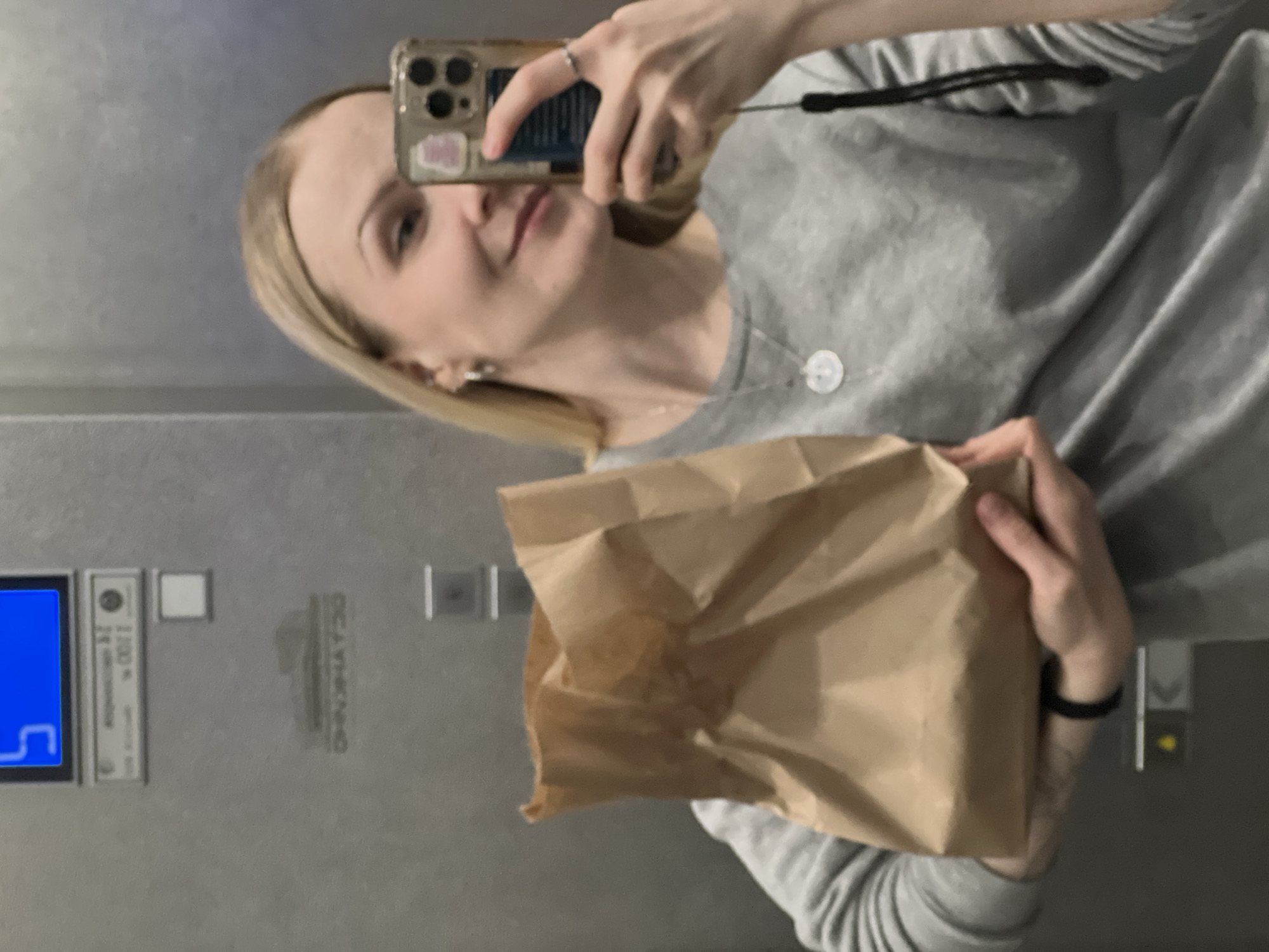 A young blonde woman is depicted in a mirror selfie, seen from the chest up within what appears to be an elevator. She holds a smartphone, visible in a clear case with a sticker, using it to capture her reflection. She is wearing a gray long-sleeved top and a simple necklace with a small, round white pendant. A silver ring is visible on her left ring finger, and a black wristband is on her right wrist. In her hands, she cradles a crumpled brown paper bag, possibly containing food or goods.
The background shows the interior of an elevator. To the left, an elevator control panel is visible, featuring a bright blue digital display showing part of a number, possibly "5". Below this, text specifies the elevator's capacity: "2100 кг" (2100 kg) and "24 человека" (24 people). Reflected on the wall or mirror behind her, the Russian word "ОГНЕТУШИТЕЛЬ" (Fire extinguisher) is visible, appearing mirrored. The elevator walls are a metallic, gray color. The woman has a slight, pleasant smile, suggesting a neutral or content mood. The overall image is rotated ninety degrees counter-clockwise.