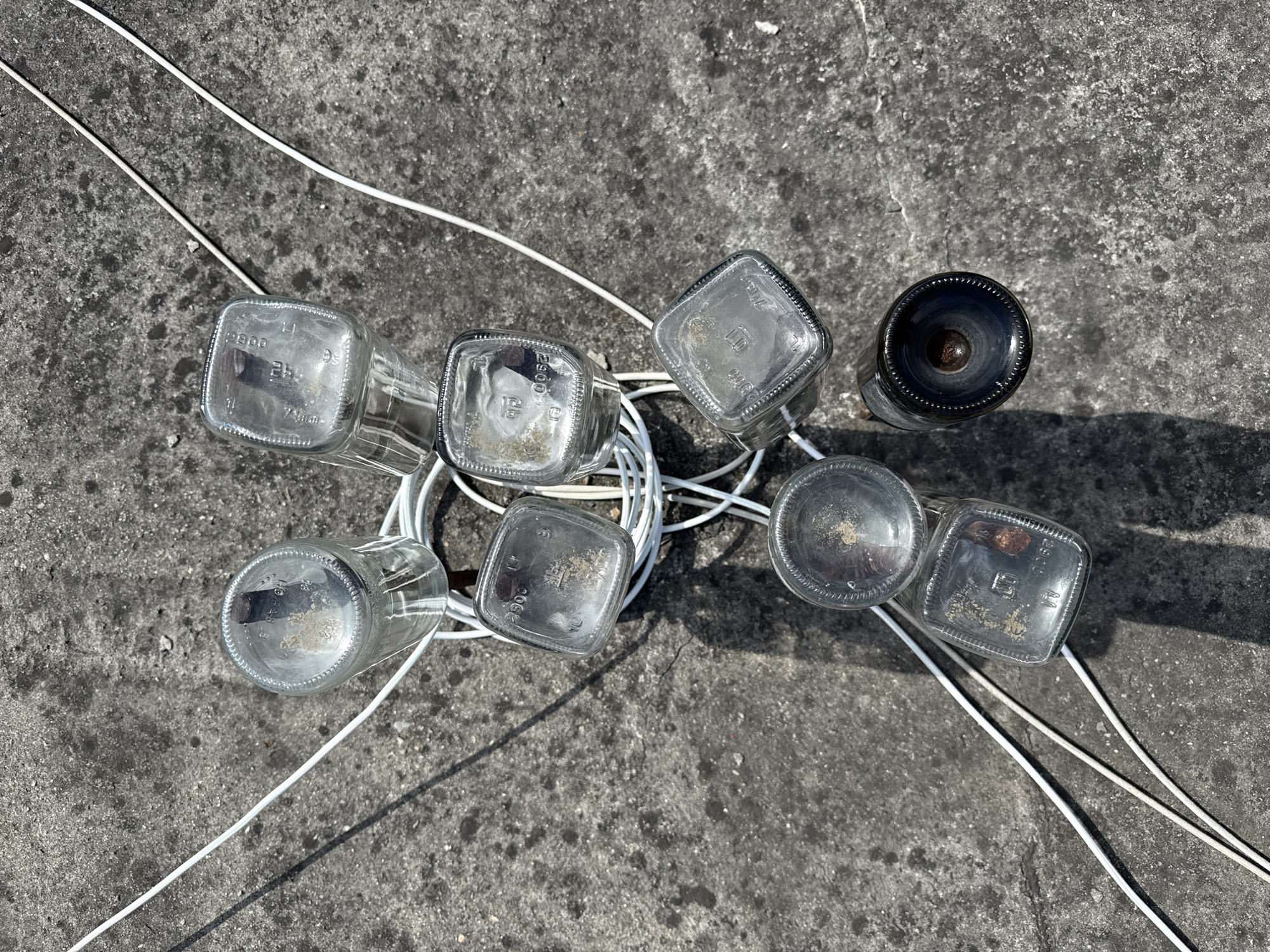 The image shows a collection of eight bottles and a coil of white cable lying on a gray concrete surface. Seven of the bottles appear to be rectangular or square in shape and made of clear glass, while the eighth bottle is cylindrical and made of dark glass. The bottles are positioned at different angles, some overlapping each other. The coil of white cable is intertwined among the bottles. The scene is well-lit, with shadows cast on the concrete surface, indicating a bright, sunny day. The location, Sochi, Russia, is not discernible from the image.