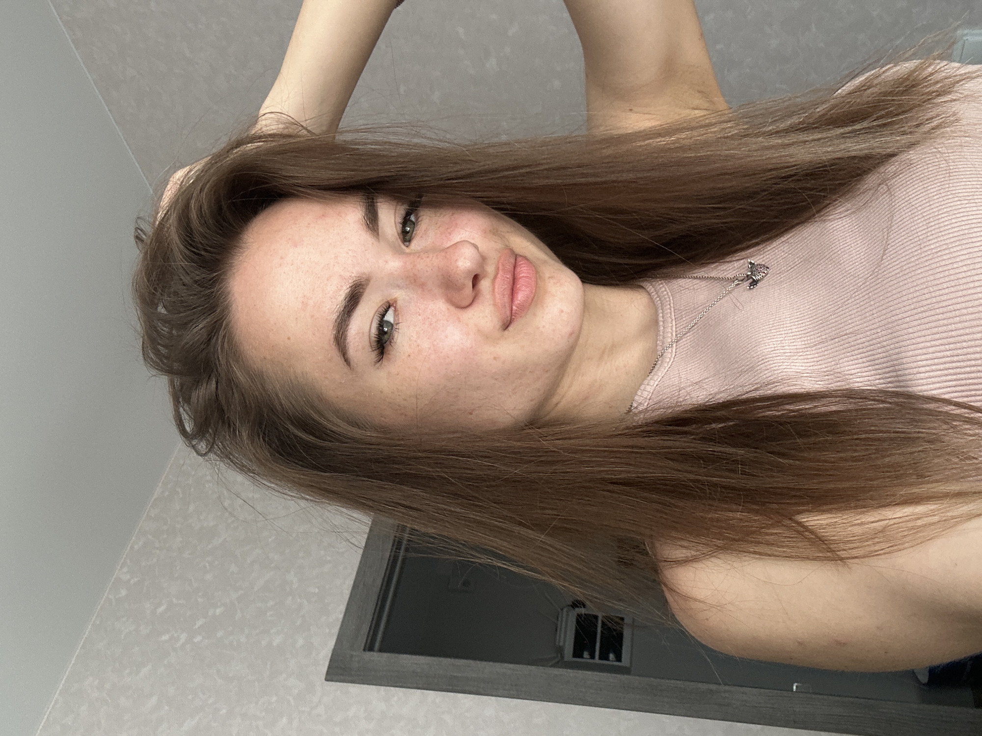 A young woman with long brown hair and green eyes is pictured indoors, facing the camera with her right hand tucked into her hair. She is wearing a light pink ribbed tank top and a silver necklace with a small pendant. The background consists of a textured wallpaper and a partially visible doorway with a light gray frame. The lighting appears to be natural, coming from the front and slightly from the left.