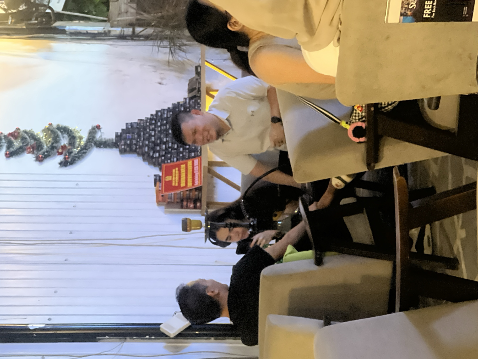 The scene depicts an outdoor area in Đà Nẵng, Vietnam. Several people are seated at tables. One man, dressed in a white shirt, is bending over, possibly interacting with a dog, based on the black fur visible near his hands. A woman with long dark hair is also looking down, perhaps also at the dog. A man wearing a black shirt is visible seated at a nearby table. In the background, a makeshift Christmas tree assembled from cans or boxes and decorated with garlands is visible against a white corrugated metal wall. A red and yellow sign is located near the makeshift tree. Tables and chairs surround the people. A sign offering "FREE" something is visible in the upper right corner of the image.