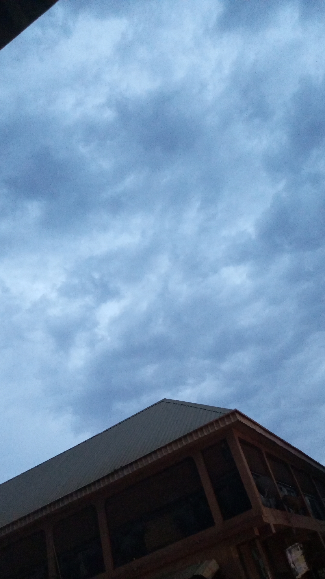 This image captures an exterior view of a building against a dramatic, overcast sky. The sky dominates the upper two-thirds of the frame, filled with dense, blue-grey clouds that suggest a cloudy day, possibly early morning or late evening due to the muted light.
The building, located in the lower right portion of the image, is angled upwards from the viewer's perspective. It features a dark, corrugated metal roof with visible ridges. Below the roof, the structure appears to be constructed from dark wood, showcasing beams and framework. Dark, recessed areas, possibly windows or open verandas, are visible along the building's side, reflecting minimal light. The overall color scheme is predominantly shades of blue, grey, and dark brown, creating a subdued atmosphere.
No people or specific activities are depicted, presenting a static architectural scene in Sabon Tasha, Nigeria. There is no visible text in the image.