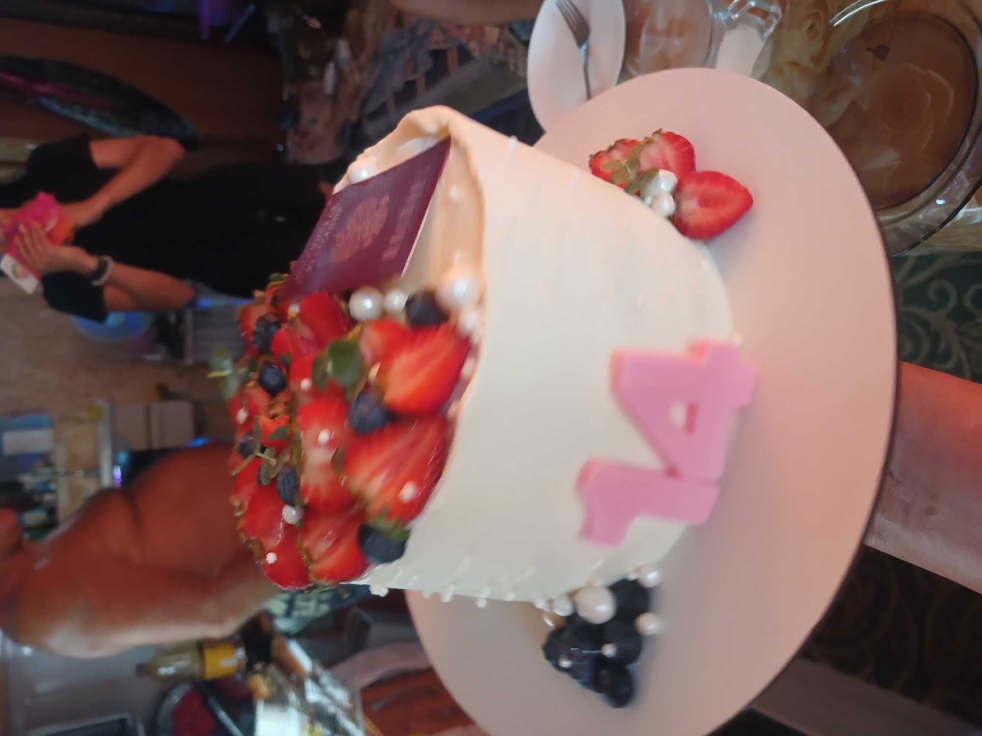 The image shows a cake decorated with strawberries, blueberries, and pearls. Two pink numbers, "14", are attached to the side of the cake. A passport is stuck into the top of the cake. In the background, a person in a black shirt is holding a pink object. The scene appears to be indoors, possibly a restaurant or a home. The overall impression is that of a celebration. The image is taken from a slightly high angle, looking down at the cake. The lighting is warm and appears to be artificial.