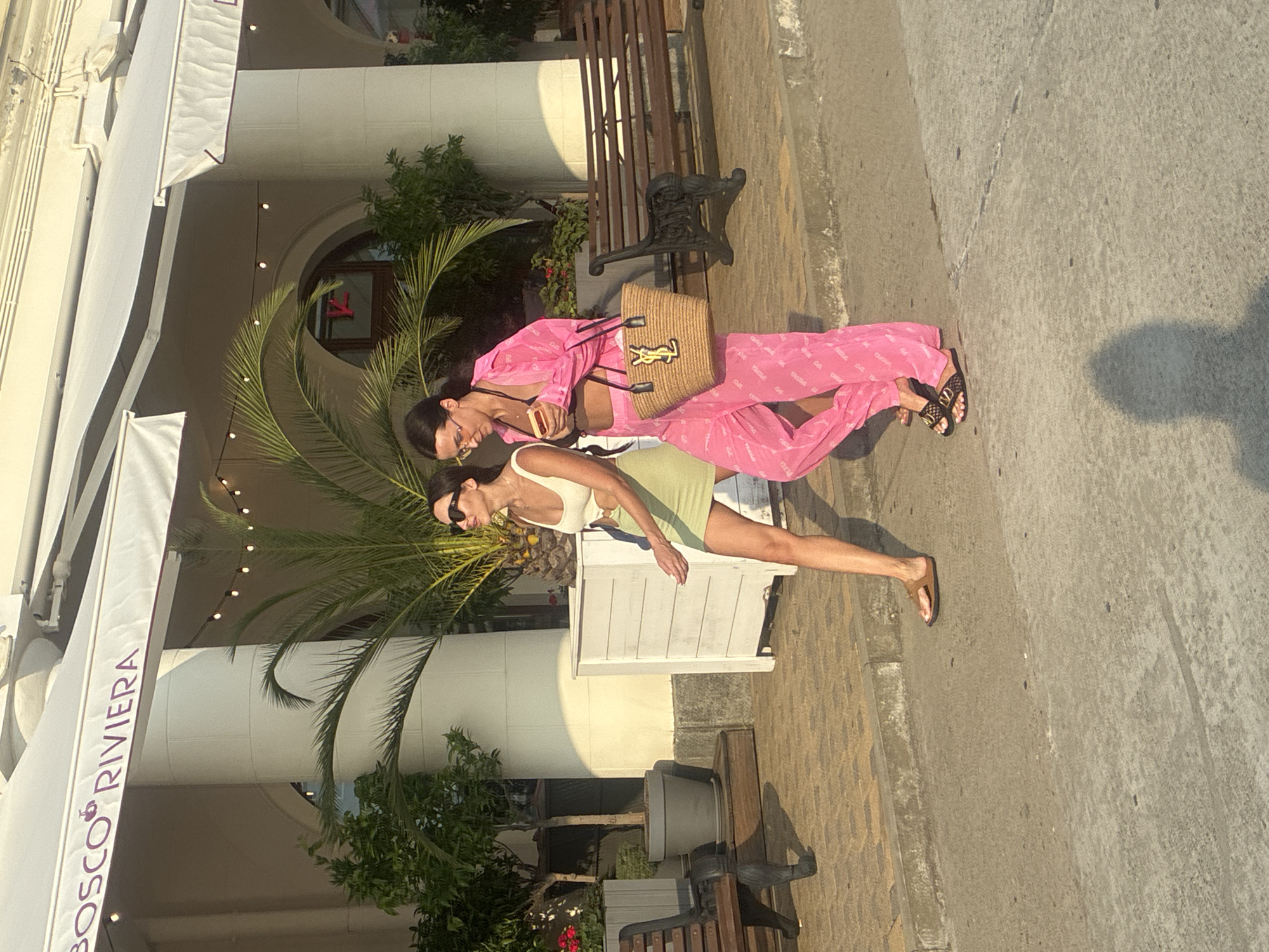 In Sochi, Russia, two women walk along a paved promenade in front of a building with awnings. The woman on the left wears a light green dress and brown sandals, while the woman on the right is dressed in a bright pink matching set of a shirt and pants, carrying a woven straw bag with a black logo. They both wear sunglasses. The building features white columns and a sign that reads "BOSCO RIVIERA." Palm trees and other greenery are visible, as well as wooden benches. The ground is a mix of patterned paving stones and a concrete surface. The scene is illuminated by sunlight, casting shadows on the ground.