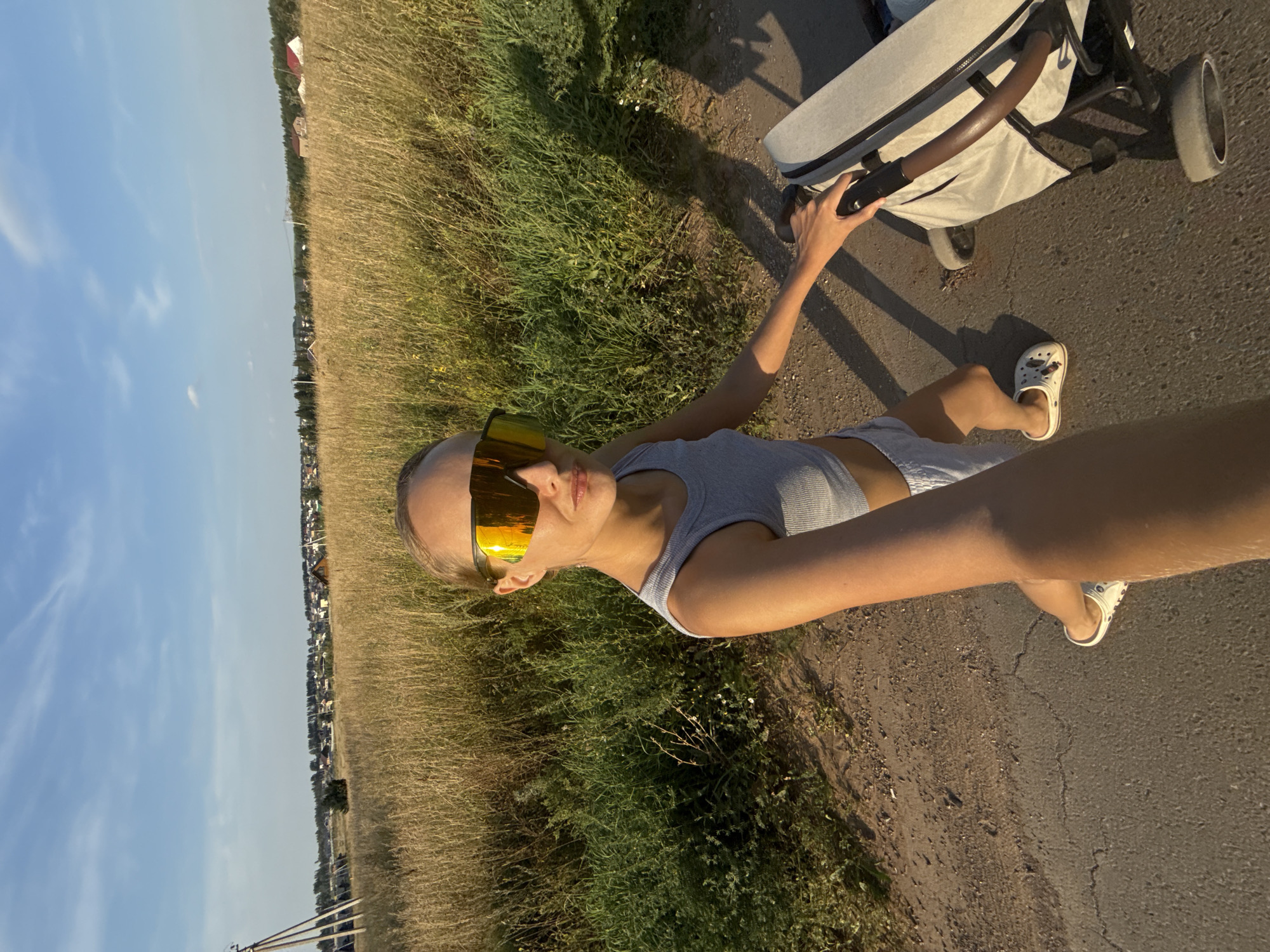 A woman stands on a dirt path in a rural area, possibly in Podshivalovo, Russia, holding the handle of a baby stroller. She is wearing a grey crop top, grey shorts, and white clogs. She has large, reflective sunglasses on. The background consists of a dry, grassy field with a few scattered trees and houses in the distance. The sky is blue with some clouds. The lighting suggests it is late afternoon or early evening.