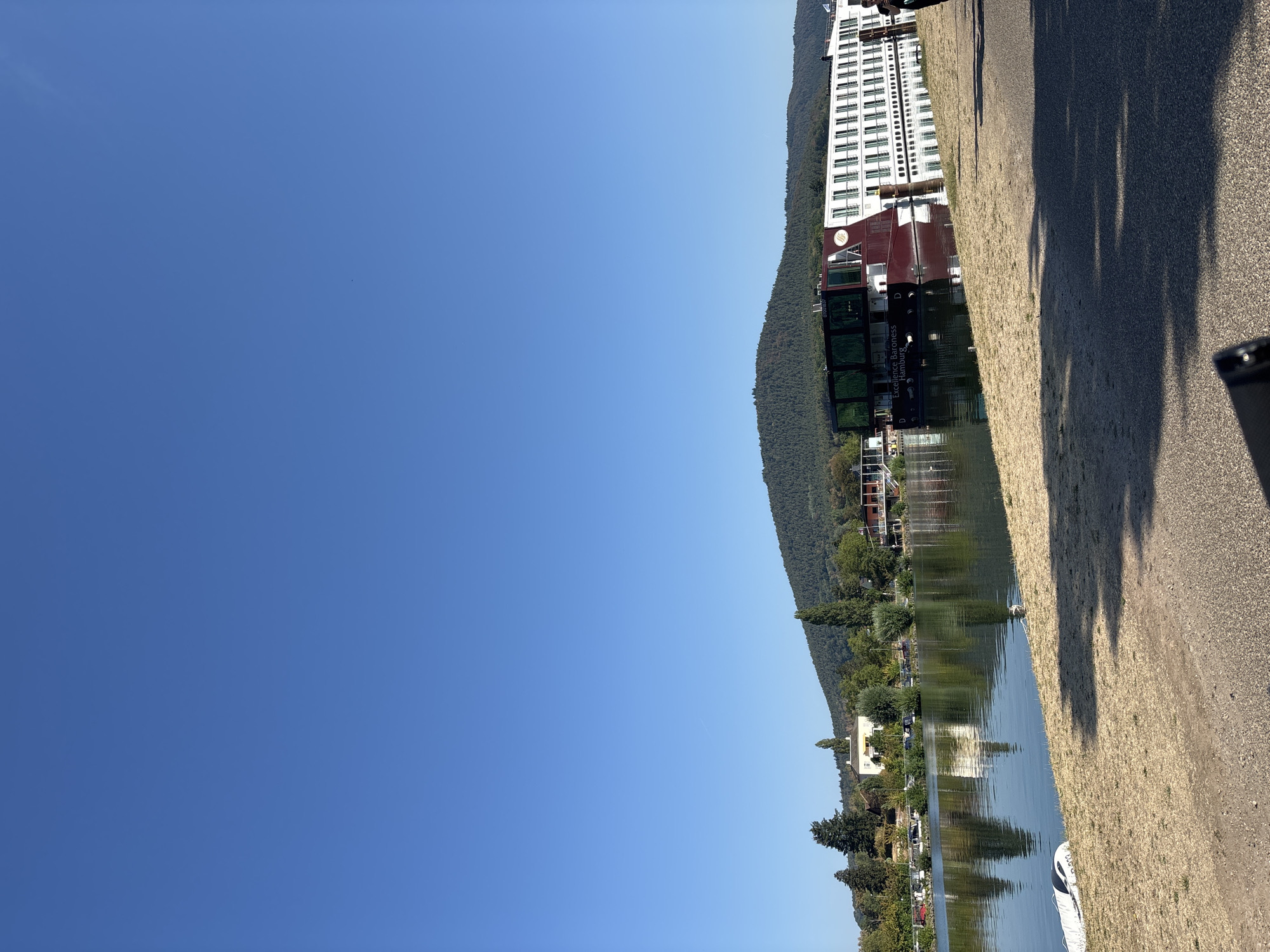 This is a vertical, eye-level, outdoor photograph taken on a clear, sunny day in Miltenberg, Germany. In the foreground, a paved path runs diagonally from the bottom right to the middle of the frame, casting dappled shadows from unseen trees. To the left of the path is a grassy bank sloping down to the water's edge. A deflated inflatable boat lies on the bank.
The middle ground is dominated by a wide, calm river, reflecting the clear blue sky and the objects on its banks. A large, white and maroon river cruise ship, identified by the text "Excellence Baroness Hamburg" on its hull, is docked along the right bank. The ship has multiple decks with rows of windows and is brightly lit by the sun. Further to the left along the riverbank, a line of trees, houses, and other buildings are visible, with a green, forested hill rising behind them.
The background is a vast expanse of clear blue sky. The overall scene depicts a peaceful waterside setting with a prominent cruise ship docked in Miltenberg, Germany.
