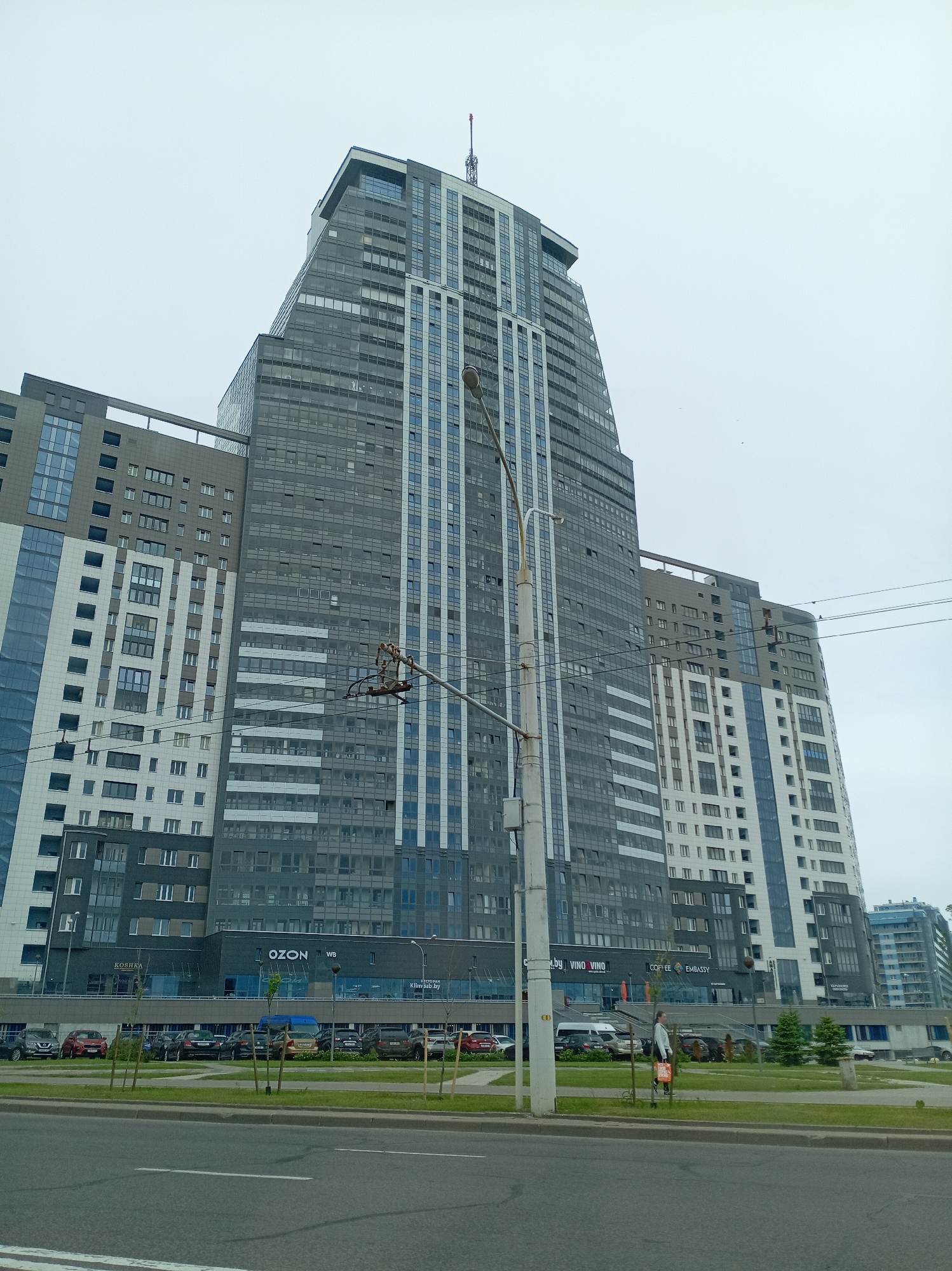 The image depicts a tall, modern building in an urban setting in the city of Unknown, Unknown. The building is primarily gray, with sections of white and blue. It has a tiered, sloping top. At the base of the building are several businesses, including "OZON," "KOSHKA," "VINO VINO," "COFFEE," and "EMBASSY." A parking lot is visible in front of the building, filled with various cars. In the foreground, there is a road and a strip of grass with trees. A person is walking on the sidewalk along the grass strip. A light pole with complex wiring is also prominent in the foreground. The sky is overcast.
