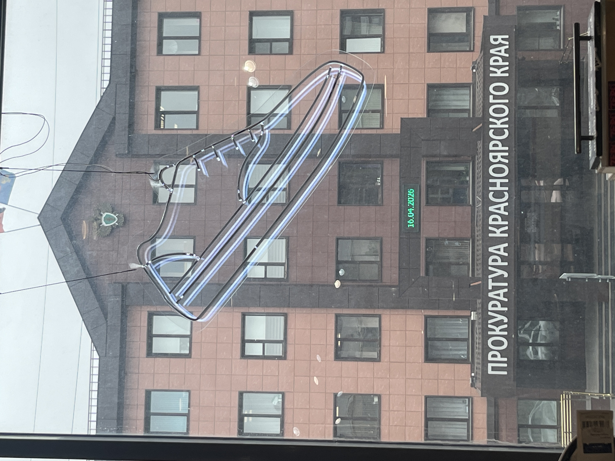 The image shows a building in Krasnoyarsk, Russia, which houses the Prosecutor's Office of Krasnoyarsk Krai. The building is a multi-story structure with a facade composed of square-shaped tiles in a reddish-brown color. The windows are rectangular and regularly spaced. A neon light in the shape of a shoe hangs on the outside of the building. A sign with the words "ПРОКУРАТУРА КРАСНОЯРСКОГО КРАЯ" (Prosecutor's Office of Krasnoyarsk Krai) runs vertically alongside the building's entrance. A digital display next to the entrance reads "10.04.2026." Wires run from the building towards the left side of the image. The photograph appears to have been taken from inside a vehicle.