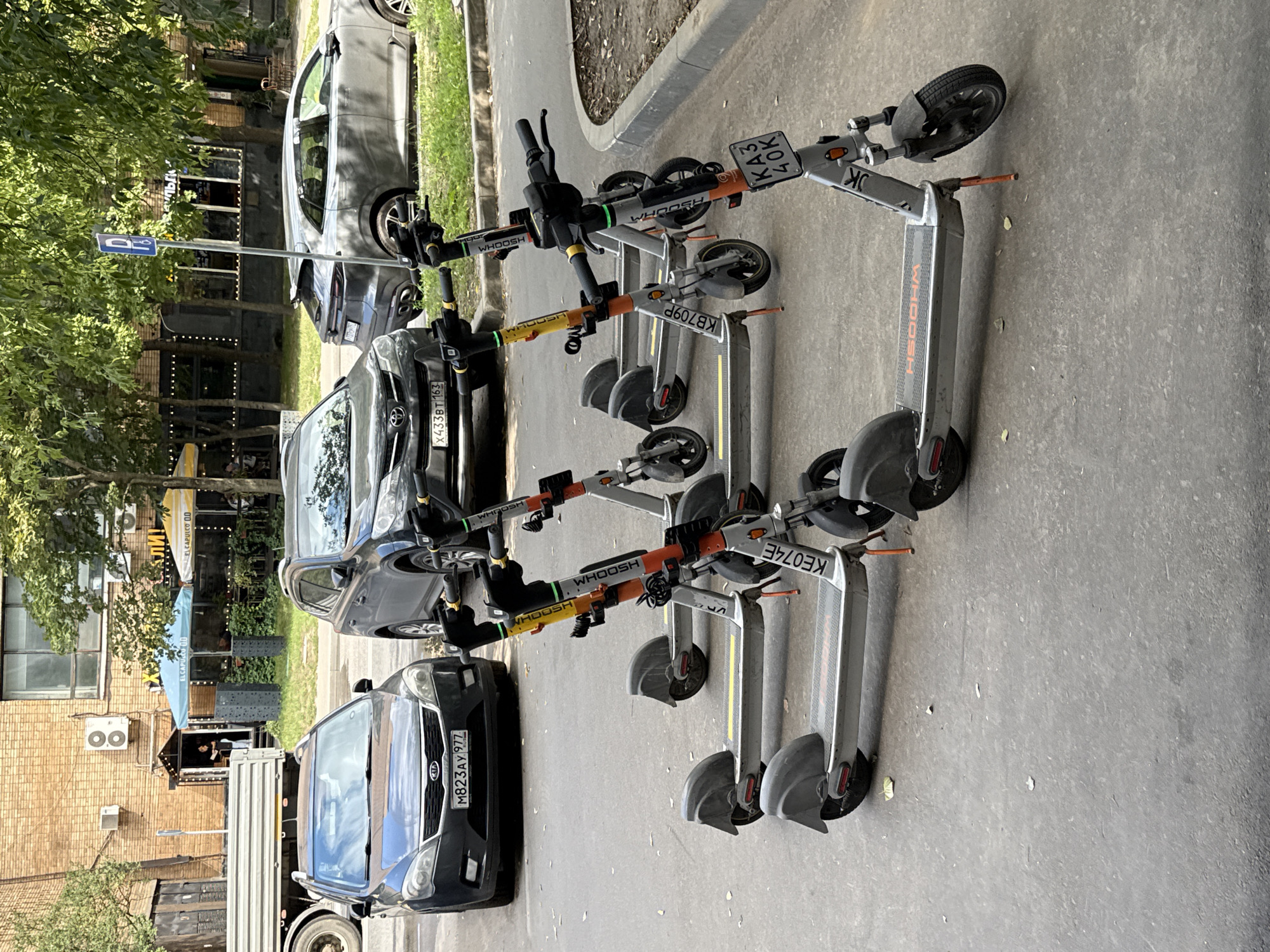In Moscow, Russia, on a sunny day, a line of four electric scooters is parked on a paved surface near a curb. The scooters are predominantly grey and silver, with orange and yellow accents. Each scooter has its brand name, "WHOOSH," visible on its deck. Three of the scooters are partially obscured by others, and the front scooter displays a license plate with the characters "KA3 40K." Behind the scooters, two cars are parked on the street: a dark grey Kia on the left, with a license plate "M823AY 977," and a silver SUV on the right. Further back, a brick building with several windows and air conditioning units stands, and trees with green foliage provide shade. In the background, two outdoor seating areas with umbrellas are visible, suggesting a cafe or restaurant. A blue sign with a wheelchair symbol is mounted on a pole to the right of the silver SUV. The scene appears to be a typical urban environment.