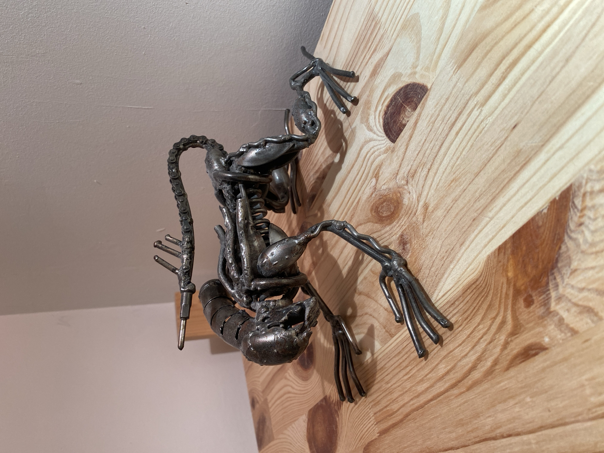 A metallic sculpture resembling a xenomorph from the movie franchise "Alien" is displayed on a wooden surface. The sculpture is dark gray and intricately detailed, with visible joints and segmented parts. It is crouched low on all fours, with its head bowed and its long, segmented tail raised. The sculpture appears to be made from recycled metal parts, such as bicycle chains and bolts. The wooden surface has a light-colored, grainy texture with prominent knots. The background is a plain, light-colored wall. The image is well-lit, with natural light casting subtle shadows. The location is not specified in Moscow, Russia.
