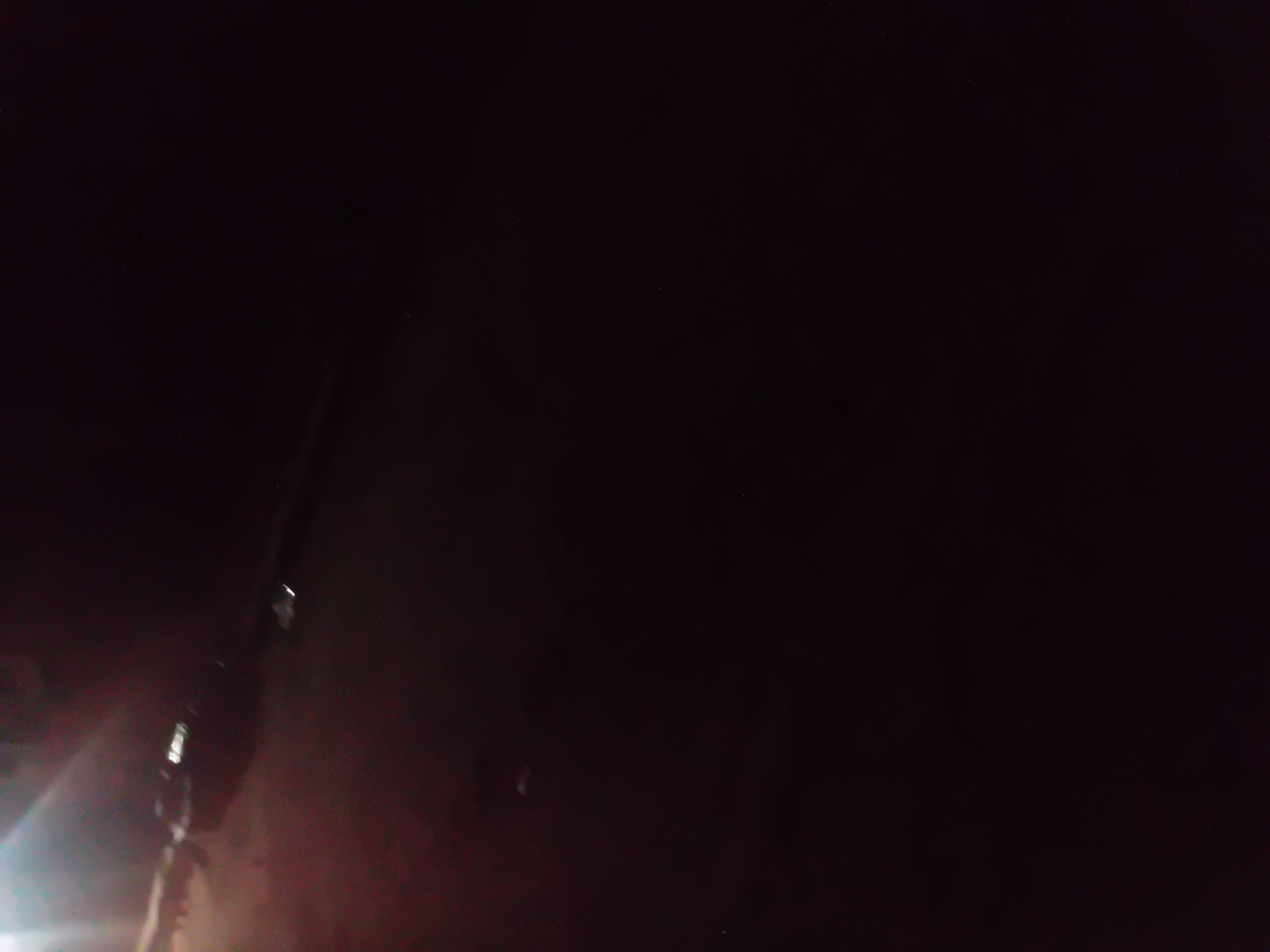 The image depicts a dimly lit, outdoor scene in Jauro Yinu, Nigeria. The predominant colors are dark, with some areas of muted reddish-brown and bright white light. In the upper left portion of the image, a bright, lens flare-like light source is visible, illuminating a portion of a dark, amorphous object or surface. This object appears to be elongated and may have reflective elements, suggesting it could be a vehicle or part of a structure. Further into the image, there are smaller, brighter white specks that could be reflections or lights from a distance. The rest of the image is largely black, with subtle variations in tone that hint at a textured or uneven surface, possibly the ground or a wall. There are no clearly discernible people or specific landmarks that allow for precise identification of the location beyond the general area of Jauro Yinu. The overall impression is of a night-time scene with minimal artificial light.