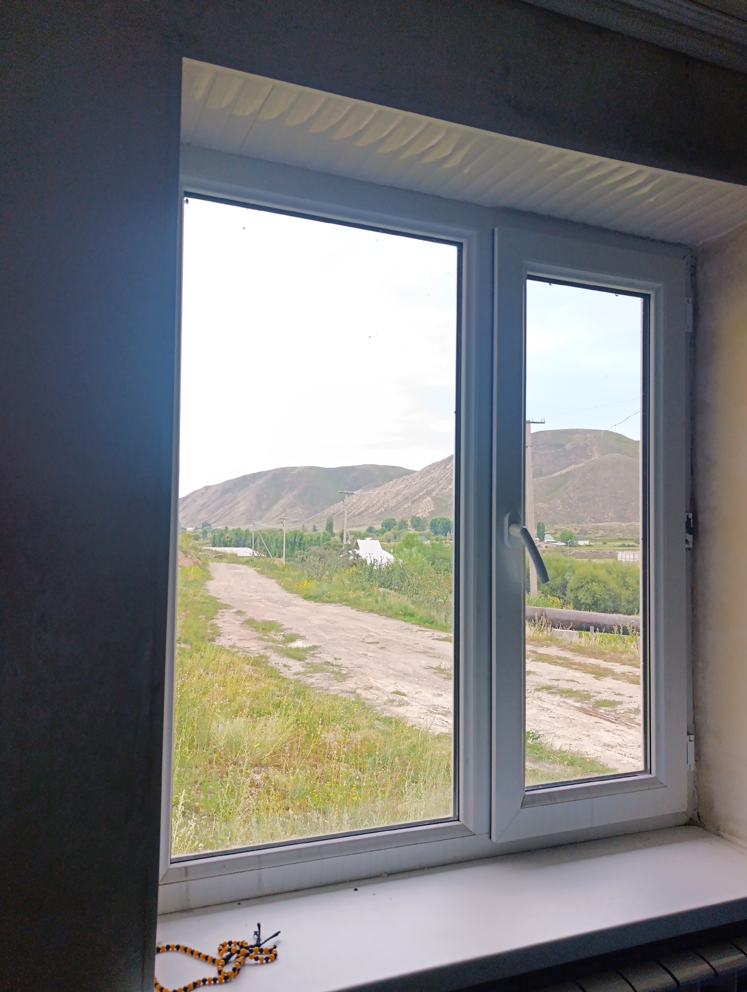 The image shows a view from a window in Kazan-Kuygan, Kyrgyzstan. The window is white and modern, with two panes. Through the window, a dirt road can be seen leading into a valley. There are green trees and hills in the background. The sky is overcast. A string of prayer beads is visible on the windowsill in the foreground.