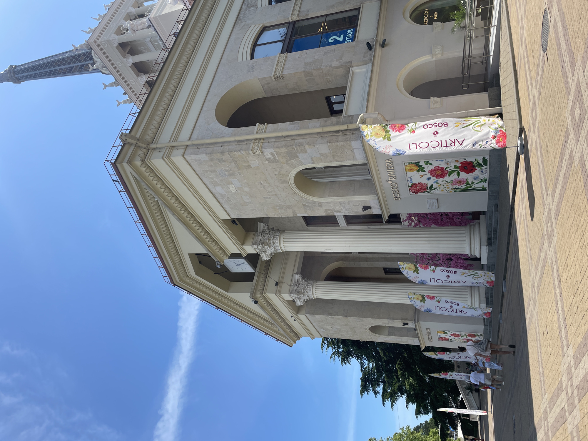 The image presents a detailed view of the Sochi Seaport building in Sochi, Russia, under a bright, clear blue sky with a few wispy white clouds. The building is a large, multi-story structure constructed from light-colored stone, featuring neoclassical or Stalinist Empire architectural elements. Its main facade showcases prominent fluted columns with ornate Corinthian capitals, supporting a pediment. A square clock with a white face and black hands is mounted beneath the pediment, centered above the columns.
To the right, a tall, dark-colored lattice spire rises from a decorative base. This base is adorned with small white sculptures, including stylized marine animals (resembling dolphin tails) and human figures. The building's exterior includes multiple arched windows and recessed areas, some fitted with dark-framed glass. A light-colored downspout is visible descending along one corner. Signage on the lower portion of the building displays "BOSCO RIVIERA," and a blue sign in an upper-story window reads "2 ЭТАЖ" (2nd FLOOR in Russian).
The foreground features a pedestrian walkway paved with light brown and reddish-brown interlocking pavers arranged in a geometric pattern. A line of five white, feather-shaped flags stands in front of the building. These flags are decorated with vibrant floral patterns (roses, peonies, and other blossoms) and display the text "ARTICOLI BOSCO" in black. Between the building's columns, large panels are similarly adorned with floral designs. To the left of the flags, a pink artificial cherry blossom tree is visible.
Approximately five to seven people are walking on the paved area, moving away from the viewer and towards the building's entrance. They are dressed in casual summer clothing, including individuals in white tops, shorts, and dresses. Dark green trees provide foliage to the left of the building. The scene is brightly lit by direct sunlight, casting distinct shadows on the pavement.