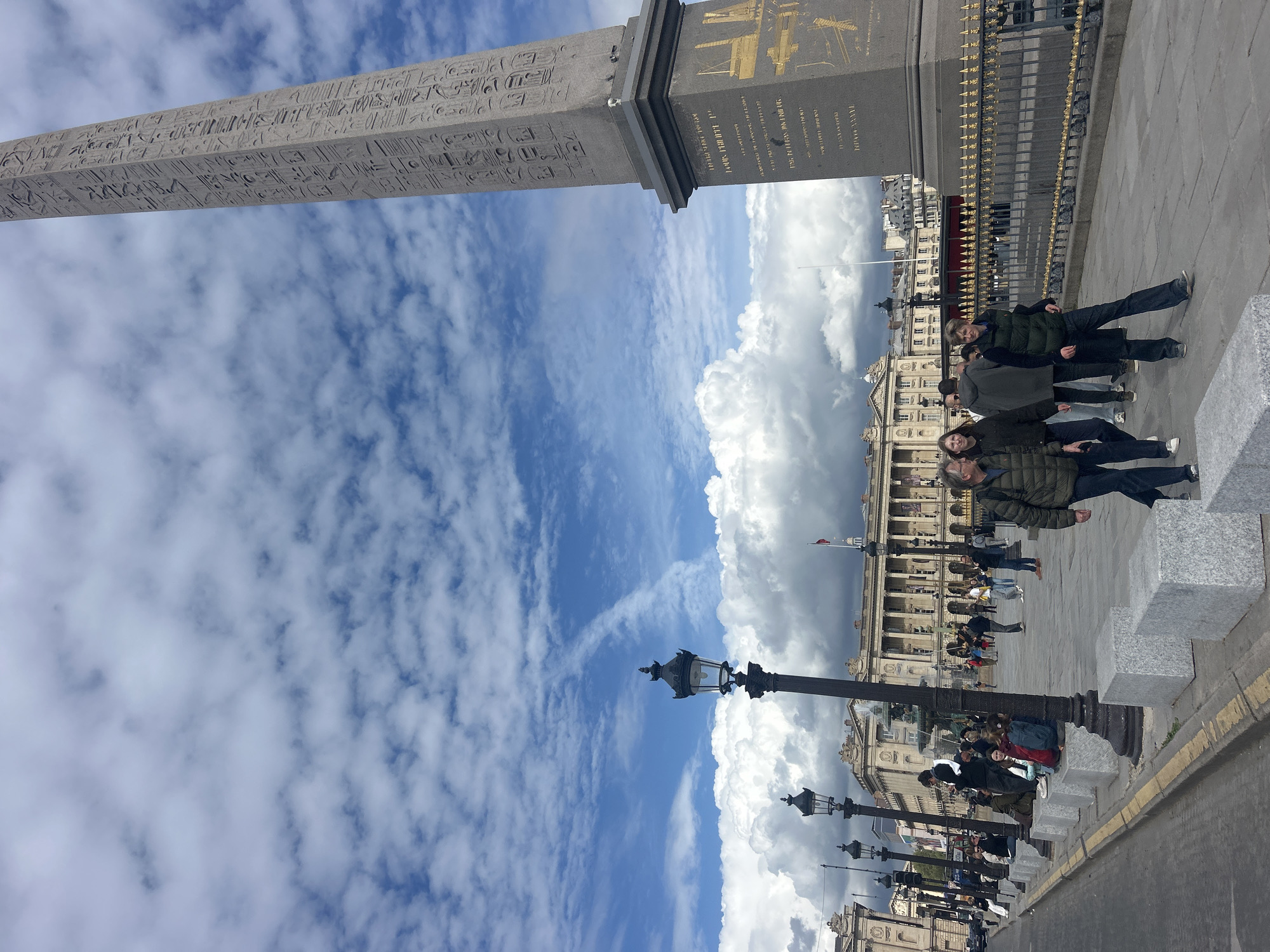 The image shows a view of the Place de la Concorde in Paris, France. The Luxor Obelisk dominates the left side of the frame, with its hieroglyphics clearly visible against the blue sky filled with scattered white clouds. To the right of the obelisk's base, a group of people stand on the paved ground, suggesting they may be tourists or locals visiting the area. The backdrop reveals the architectural details of the buildings surrounding the Place de la Concorde. Street lamps line the edge of the square.