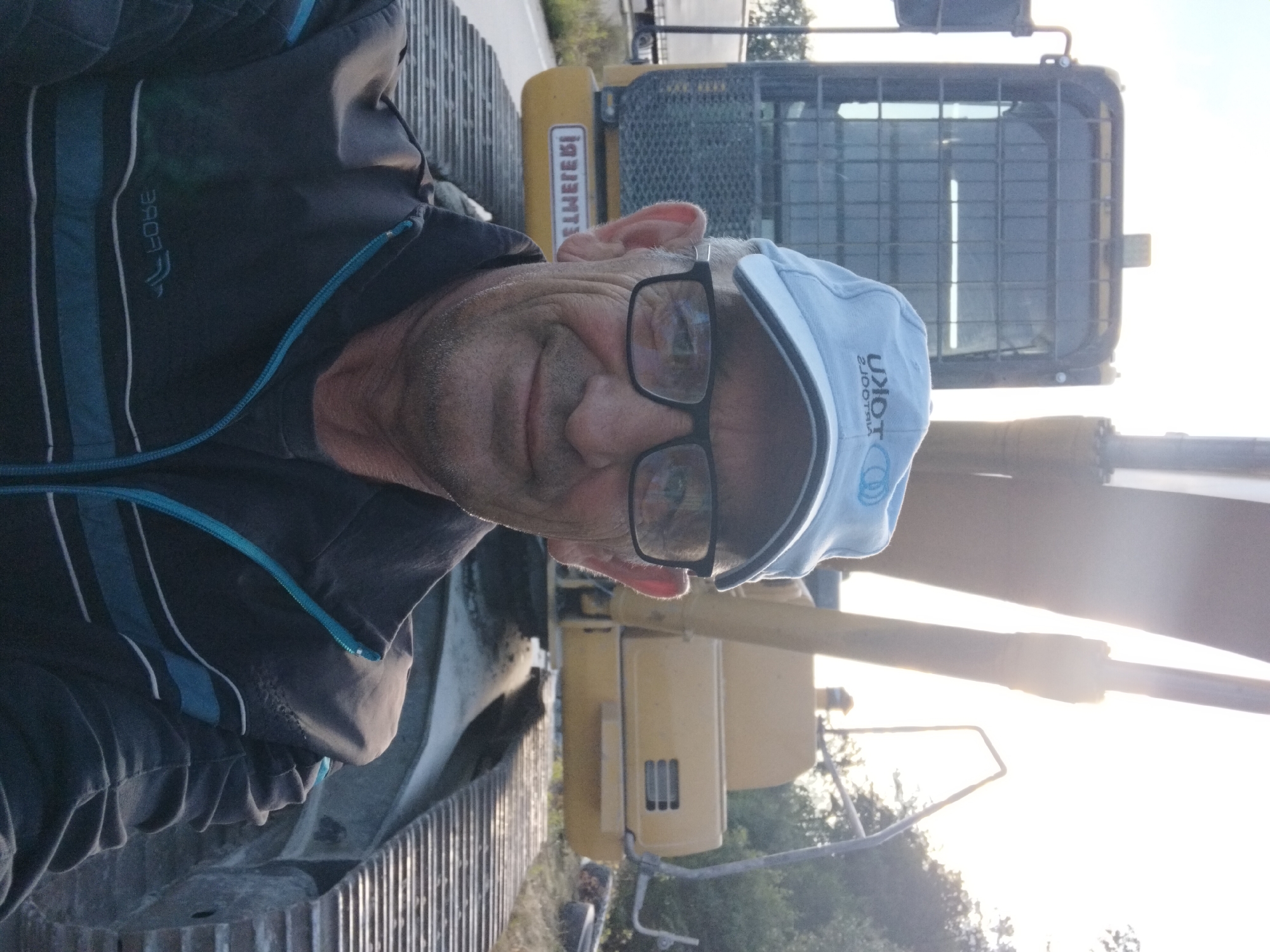 A man wearing a light blue baseball cap and eyeglasses is taking a selfie in front of a yellow excavator. He is wearing a dark jacket with blue and white stripes. The excavator has a yellow cab and large metal tracks. The background shows trees and a bright sky. The image appears to be taken in a rural or industrial area, possibly on the outskirts of Novoselytsia, Ukraine.