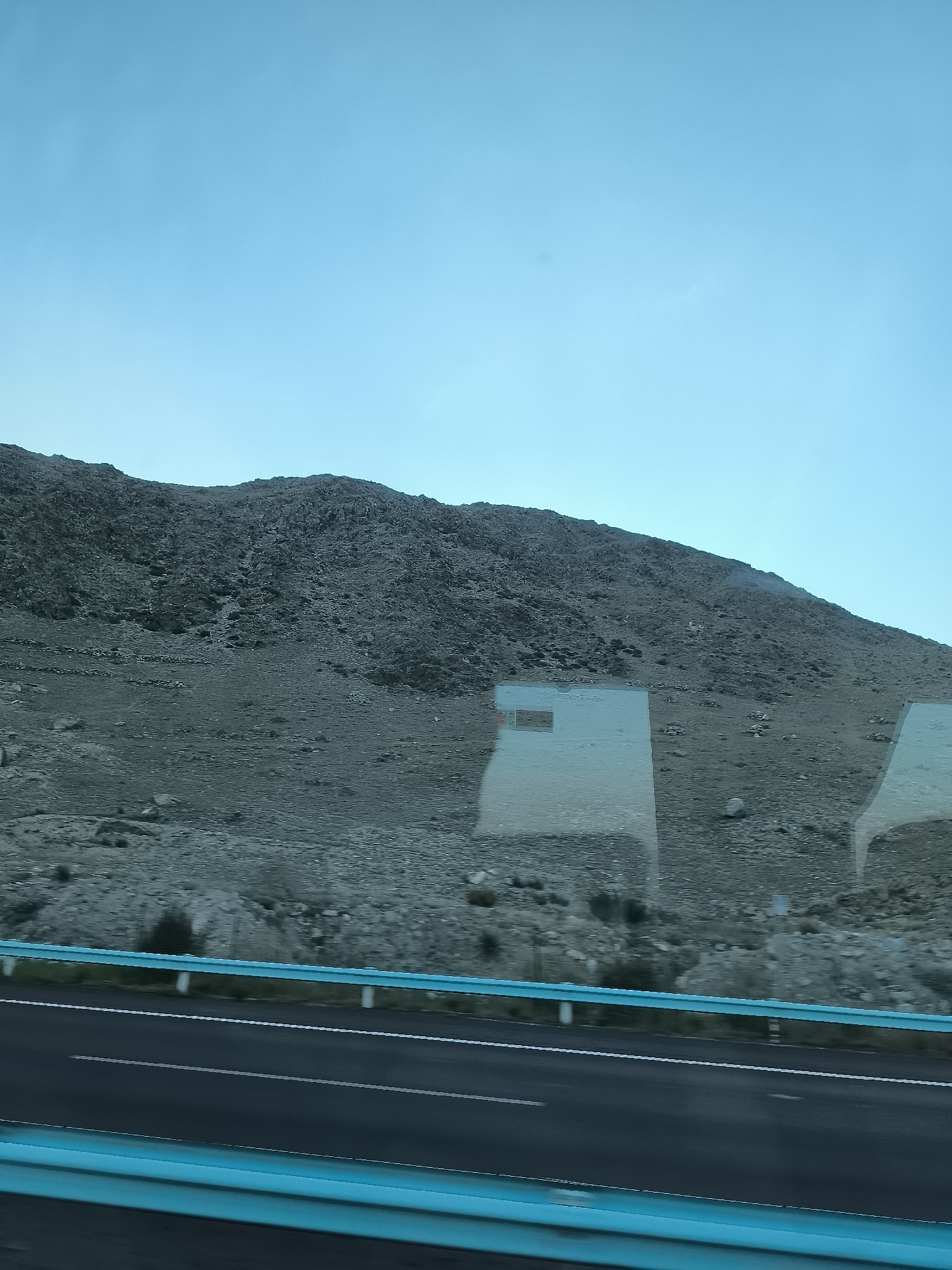 The image shows a barren, rocky hillside under a clear blue sky, viewed from a moving vehicle. The hillside is covered with small and large rocks, with sparse vegetation visible in places. A highway with two lanes and a turquoise guardrail runs along the bottom of the frame. Reflections of the vehicle's interior are visible on the windows, obscuring parts of the landscape. The scene appears to be a remote, undeveloped area, possibly a mountainous region in China, given the instruction.
