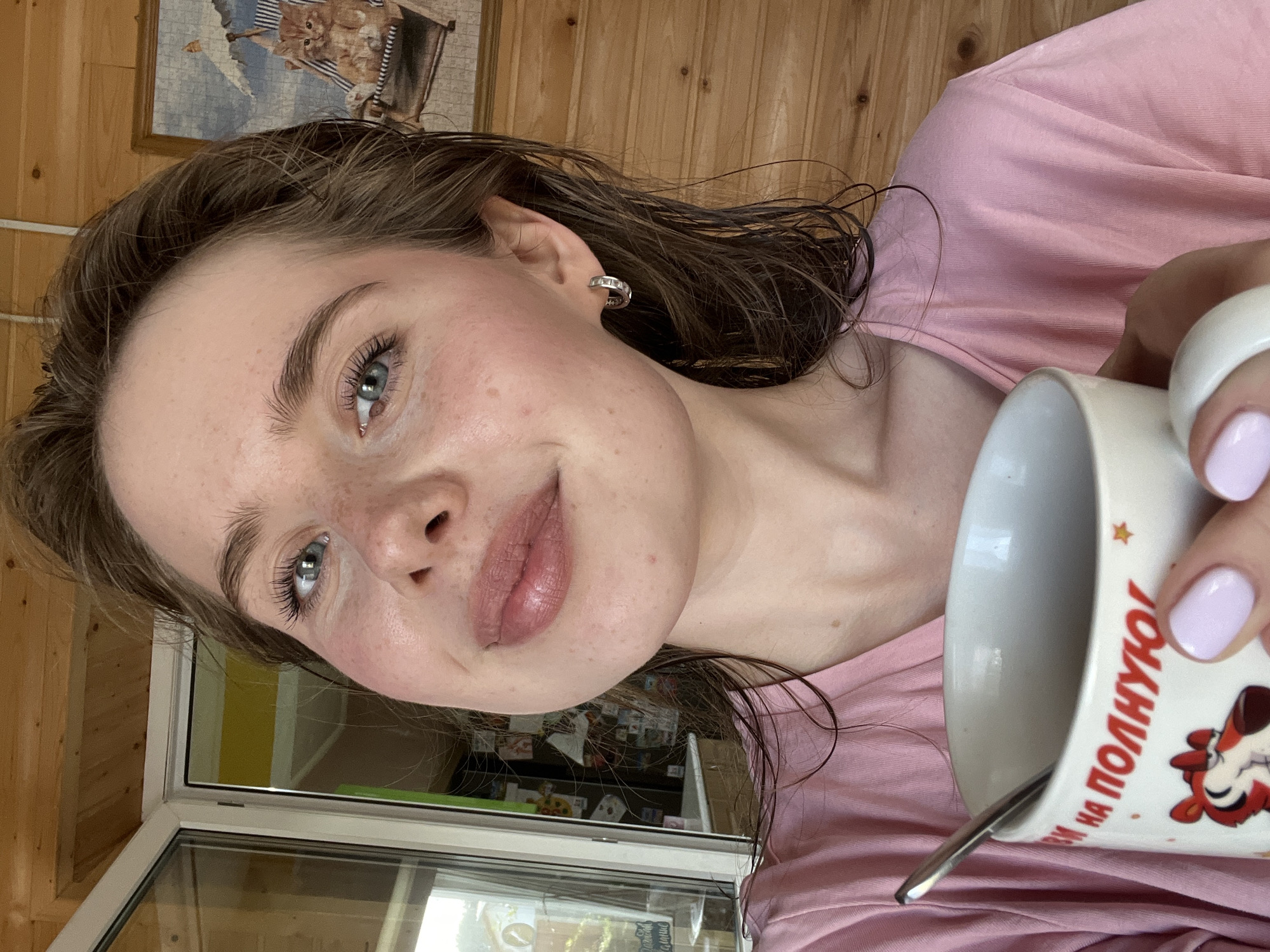 A young woman with blue eyes, light brown hair, and freckles is taking a selfie. She is wearing a pink t-shirt and holding a white mug with a spoon in it. The mug has a cartoon tiger and red Russian text on it. The background appears to be a wooden interior with a window showing an outdoor scene. The overall impression is of a casual, home-like setting. The location is the city of Chaikovsky, Russia, a city located in Perm Krai, Russia, situated on the Kama River.
