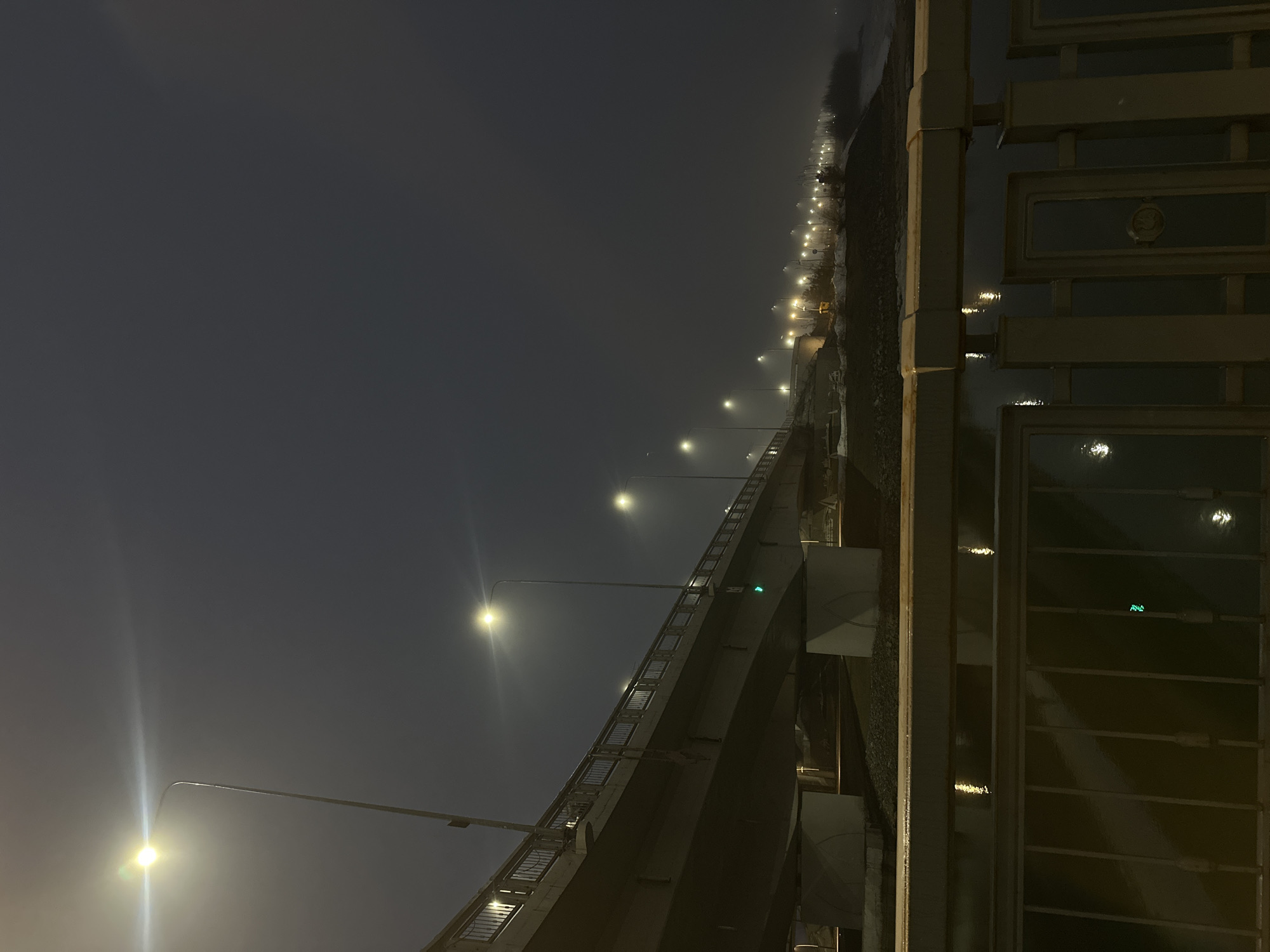 A low-angle, nighttime view of a bridge with streetlights lining its length. The photo seems to be taken from or near a railing. Streetlights stretch into the distance along the bridge. A concrete structure is below the bridge. The environment is misty or foggy. The location is most likely Kazan, Russia.