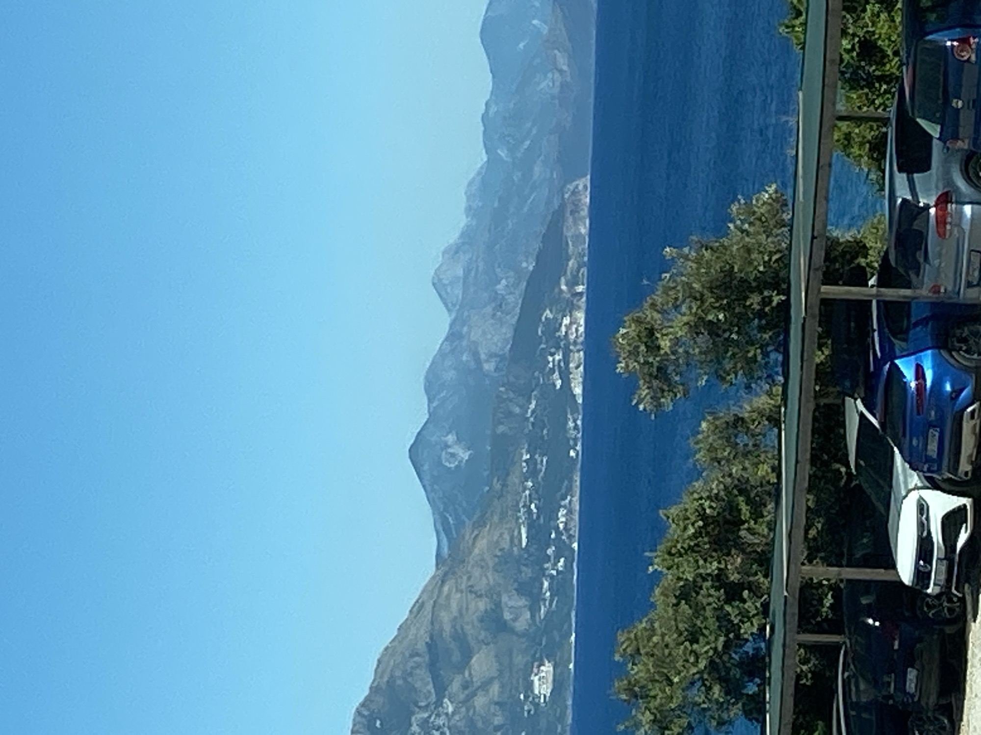 A high-angle, full shot captures a parking area in Prijevor, Montenegro, with a scenic backdrop of the Adriatic Sea and mountains. The parking lot is covered by a dark green awning supported by wooden posts, and several cars are parked beneath it. These include a dark blue SUV on the far left, a white SUV in the center, a blue hatchback to its right, and a silver hatchback further to the right. Beyond the parking lot, lush green trees provide a natural foreground to the vibrant blue sea. In the distance, a range of majestic mountains, some with white accents, dominates the horizon under a clear, bright blue sky. The scene depicts a sunny day in a coastal mountain region.