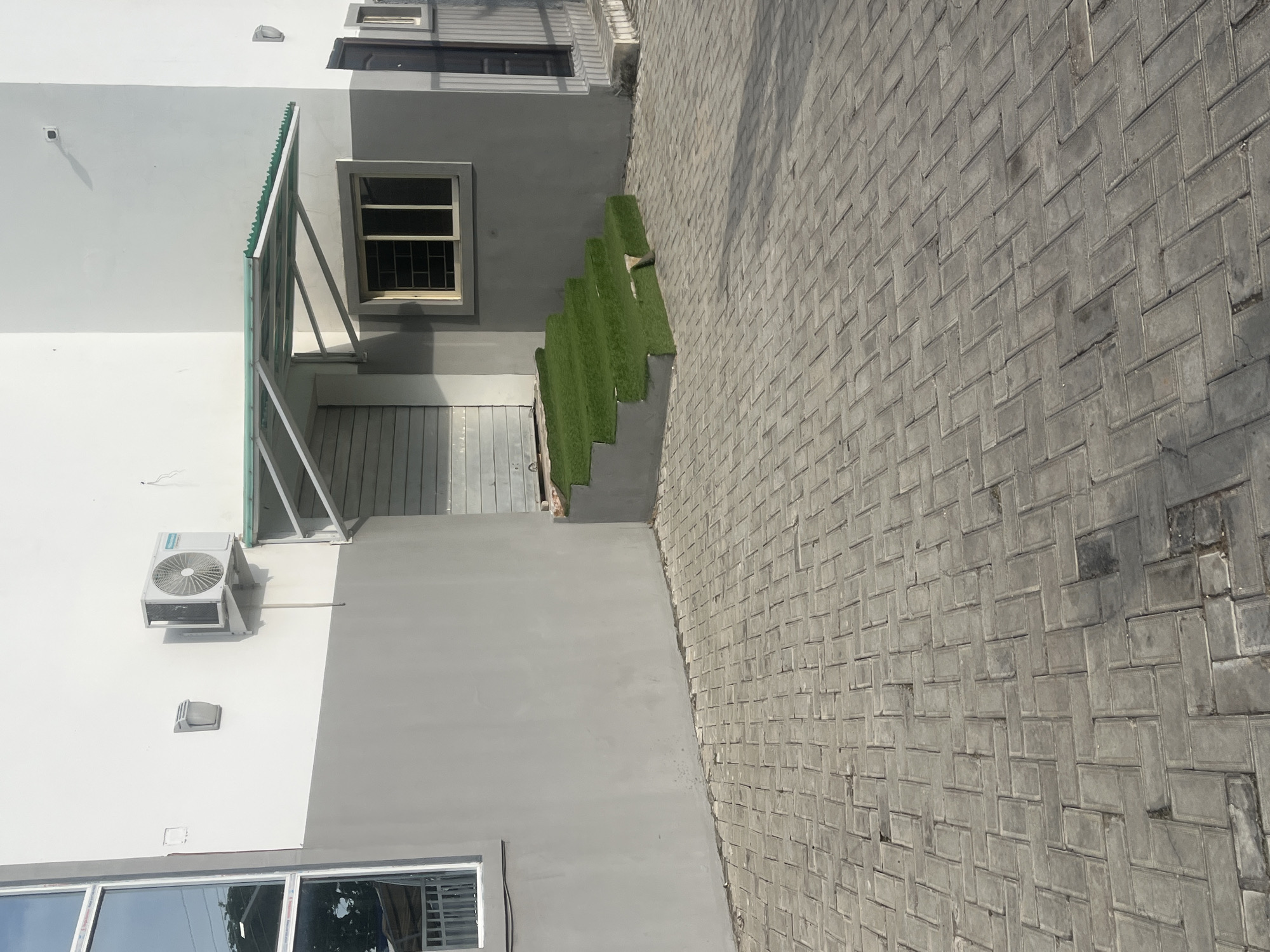 This is an outdoor, eye-level shot of a modern building facade and paved walkway. The building features a light gray lower section and a white upper section. An air conditioning unit is mounted on the white wall. To the left, a large window with a gray frame is visible. Extending from the building is a covered walkway with a green, transparent roof supported by metal beams.
Adjacent to the building, a set of stairs is covered with artificial green turf, creating a striking contrast with the surrounding concrete. The walkway is paved with rectangular gray interlocking bricks arranged in a herringbone pattern. The lighting suggests it is daytime, likely sunny, given the distinct shadows.
There are no people visible in the image. The scene appears to be a residential or commercial property exterior in Jalingo, Nigeria, characterized by its clean lines and modern design elements. There is no visible text in the image.