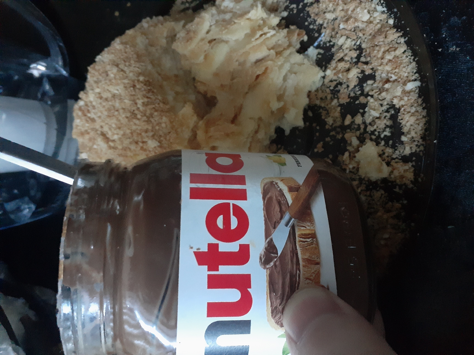 A jar of Nutella sits on a black surface next to a pile of crumbled cake. A knife with Nutella on it is held by a thumb and forefinger, reaching towards a slice of bread with Nutella on it, which is part of the label on the jar. The background is dark and out of focus. The scene is lit from above, casting shadows.