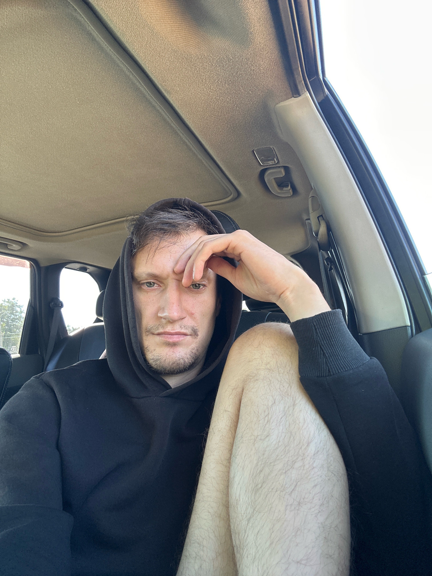 A medium shot shows a man sitting in the driver's seat of a vehicle, wearing a black hooded sweatshirt. He is looking directly at the camera with a contemplative expression, his left hand resting on his forehead. His right leg is bent and crosses in front of his left. The interior of the car is visible, including the headliner and a portion of the door panel. The scene appears to be outdoors, with sunlight suggesting a daytime setting. The city of Makiv, Ukraine, is not discernible from the visual information presented.