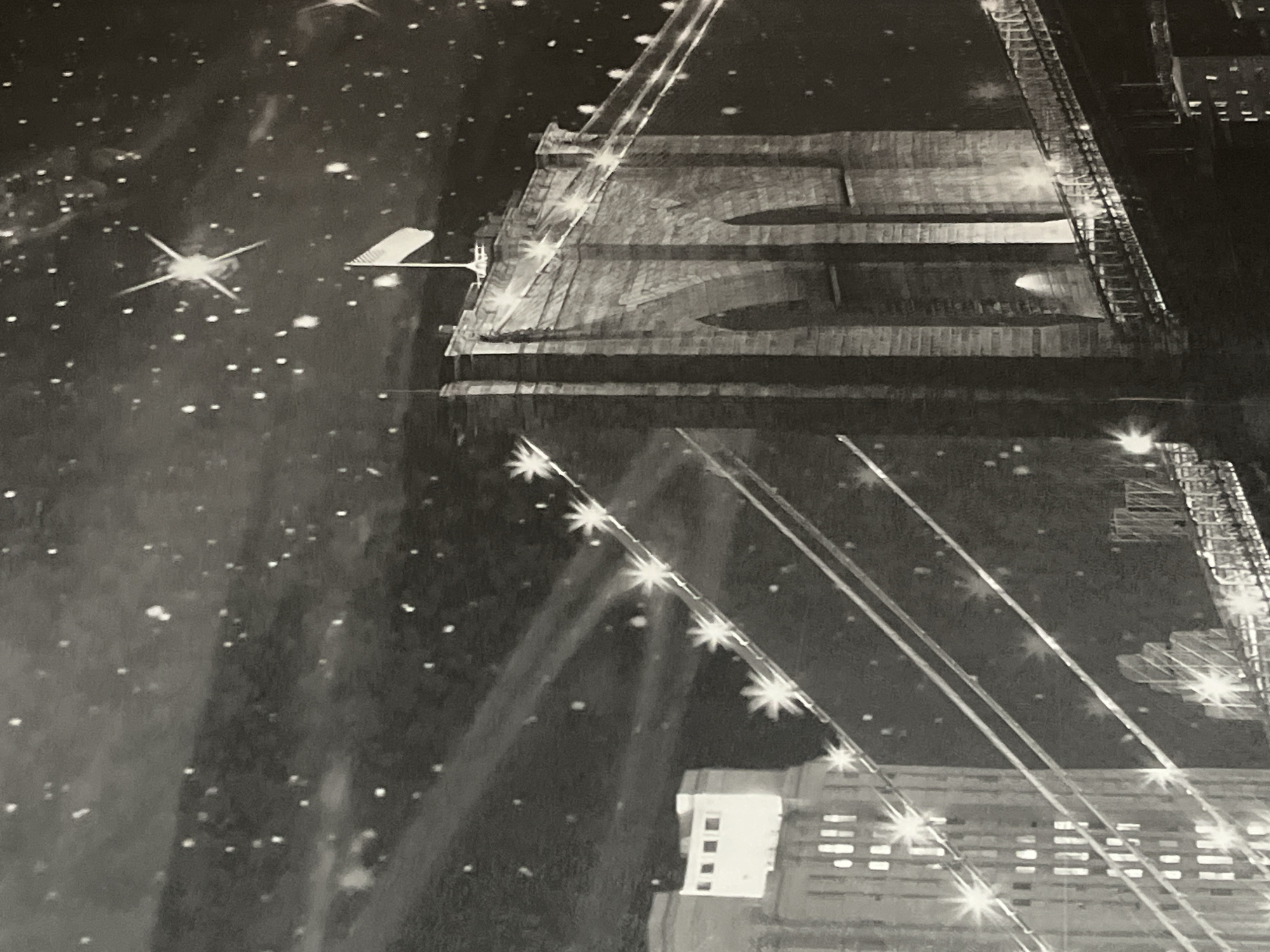 The image depicts a nighttime view of the Brooklyn Bridge in New York City, United States. The iconic bridge is illuminated, with its suspension cables and towers clearly visible against a dark sky filled with numerous stars and light streaks. A flag flies from the top of one of the bridge towers. On the left side of the image, a tall, illuminated building with multiple windows stands. The scene is rendered in black and white, giving it a classic or artistic quality. There are no people or specific actions immediately discernible in the image. The environment is urban, characterized by the bridge and the city skyline. The location is explicitly identified as the Brooklyn Bridge, New York City, not Dnipro, Ukraine.