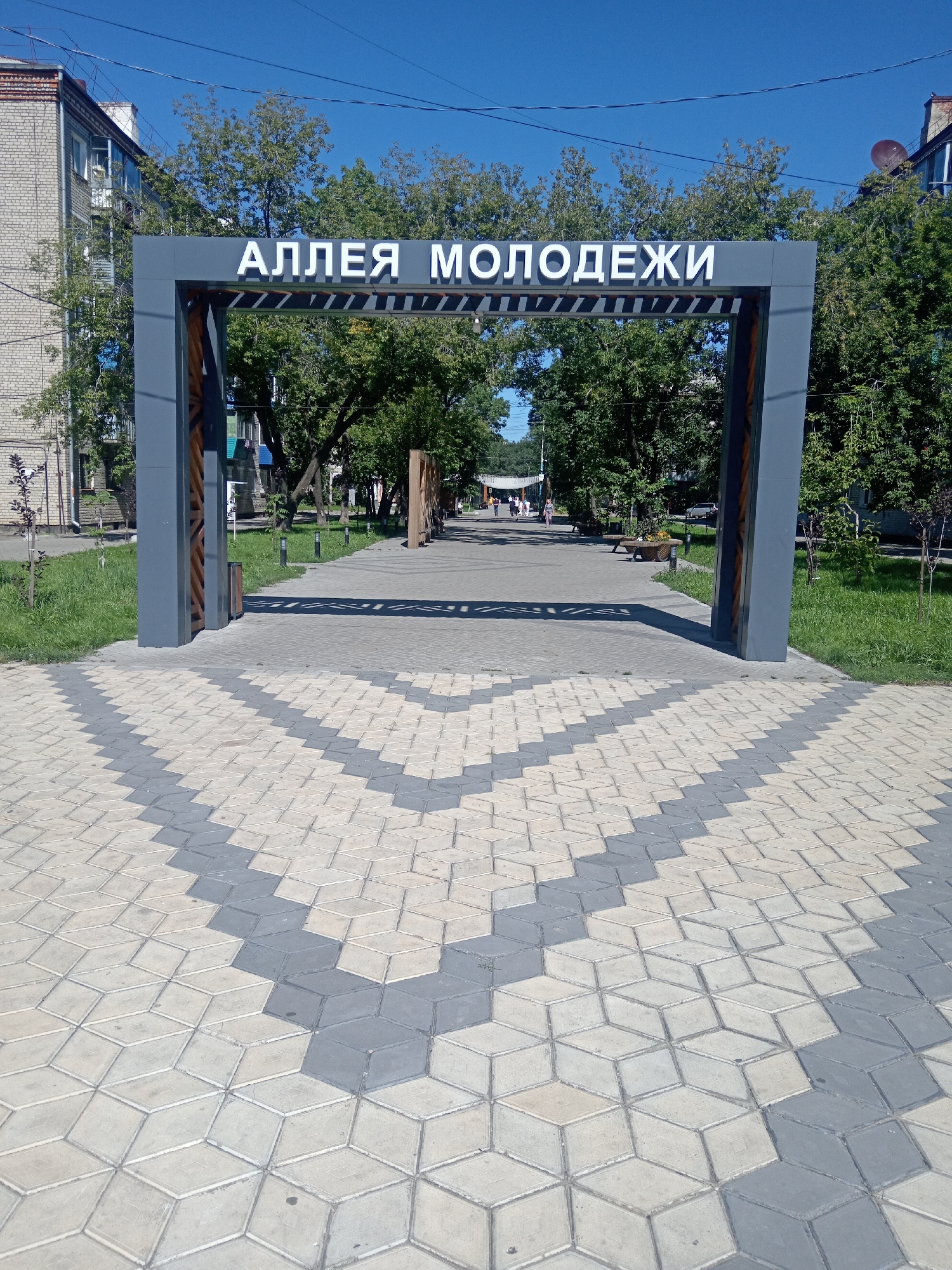 A wide-angle, eye-level shot captures an arched entryway in Belogorsk, Russia, marking the entrance to "Alleya Molodezhi" (Youth Alley). The archway, constructed of gray metal with wooden accents, frames a pathway lined with trees and buildings. The ground in the foreground is paved with geometric, interlocking tiles in shades of light gray and beige, creating a striking visual pattern that leads towards the arch. Beyond the arch, the pathway continues, with a few people walking in the distance. Benches are visible along the sides of the path, and the sky above is clear blue. The overall scene depicts a well-maintained urban public space on a sunny day.