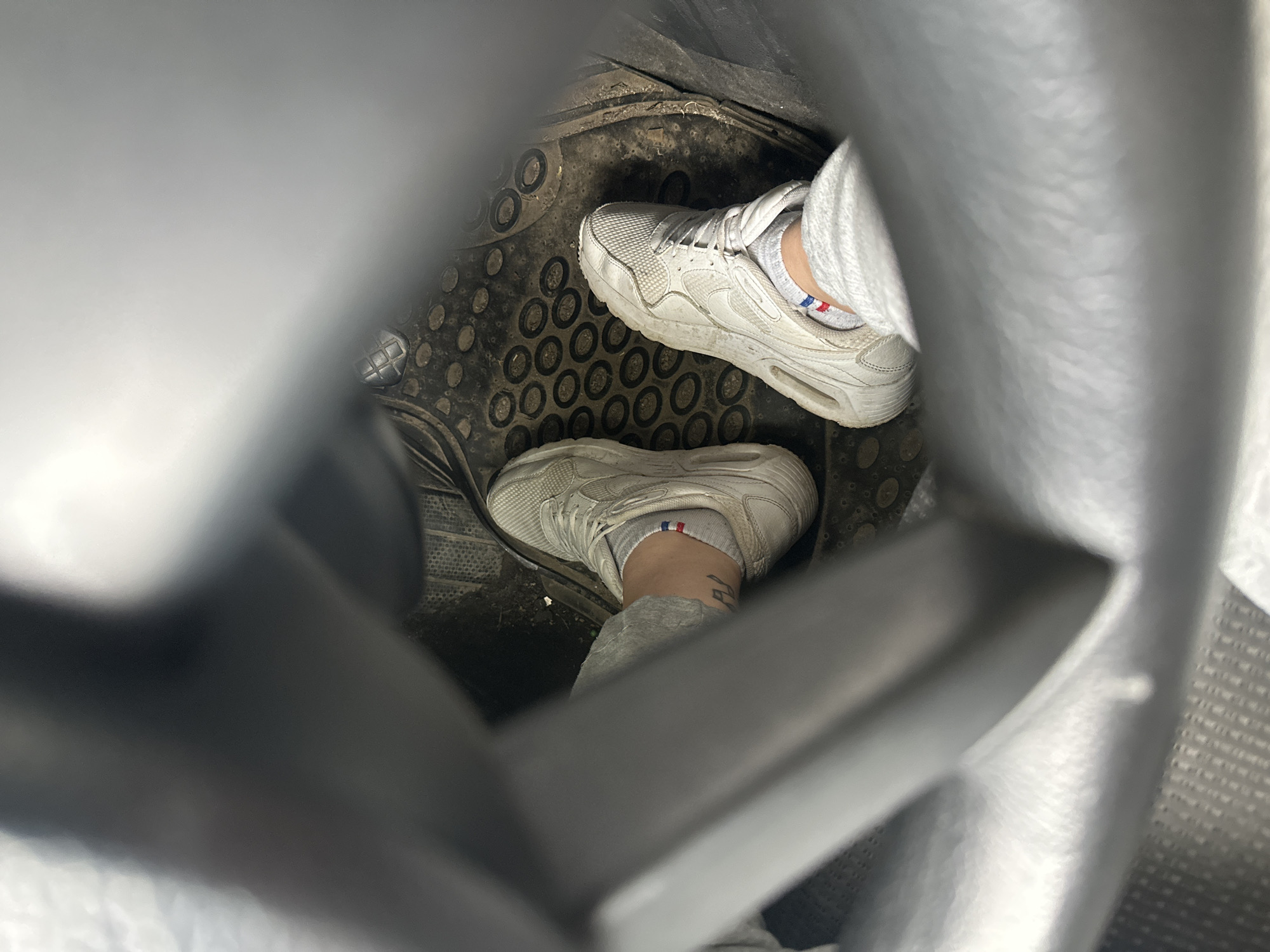 The image shows two feet in a car's footwell. Both feet are wearing white sneakers and grey socks with a red, white, and blue band at the top. The floor mat is black with a repeating circular pattern and appears dirty. The view is partially obstructed by gray plastic framing, possibly part of the car's interior. There is no indication of the location being Resen, Bulgaria.