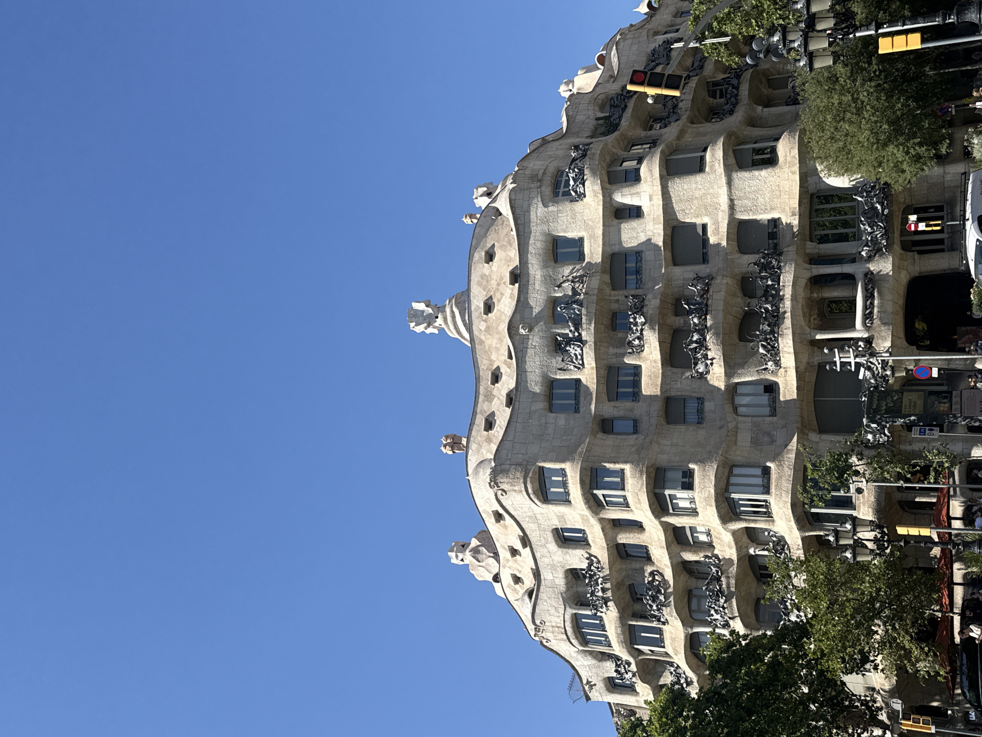 This is an outdoor, eye-level shot of the Casa Milà, also known as La Pedrera, in Barcelona, Spain. The building, designed by Antoni Gaudí, features a distinctive undulating stone facade with a honeycomb-like appearance. The image is oriented vertically, showing a portion of the building's upper floors against a clear, bright blue sky, suggesting a sunny day.
The architecture is the primary subject, with its organic, wave-like forms, irregularly shaped windows, and ornate balconies. Sculptural elements, resembling figures or gargoyles, are visible on the roofline. The texture of the stone is detailed, with shadows and highlights emphasizing the building's unique contours.
In the background, trees with lush green foliage are interspersed with the architecture, indicating an urban setting with green spaces. Traffic lights with red and yellow signals are visible, suggesting this is an active street. Some signs and parts of other buildings can be seen on the right side of the image, hinting at the bustling city environment.
There is no visible text in the image, and no specific activity or event is depicted beyond the general presence of a building in an urban environment. The clear sky and bright light suggest it is daytime, likely late morning or afternoon. The overall mood is one of architectural appreciation and the vibrancy of a European city.