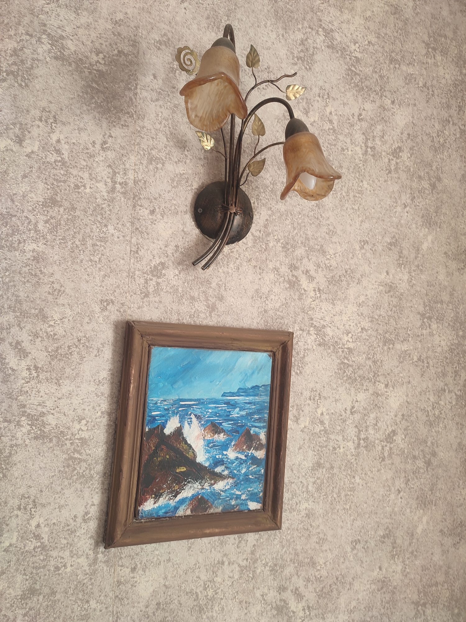 This image depicts a wall with textured wallpaper, featuring a decorative wall lamp with two amber-colored glass shades and metallic leaf accents. Below the lamp, a framed painting of a seascape is hung. The painting shows a rough sea with white waves crashing against dark, rocky shores under a bright blue sky. The style of the painting appears to be oil on canvas, with visible brushstrokes. The overall impression is one of a cozy, home interior decoration, possibly in Kriukivshchyna, Ukraine, given the location context. No people are visible in the image, and no specific time of day or weather conditions can be inferred from the scene beyond the bright sky in the painting. There is no visible text in the image.