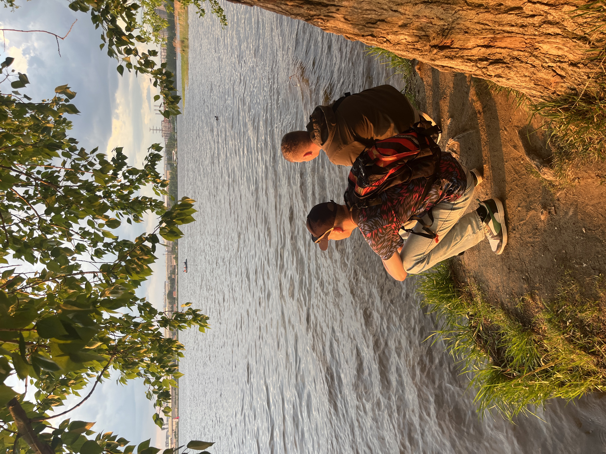 Two people are positioned at the edge of a body of water. The person closest to the camera is wearing a red and black patterned shirt, jeans, a black cap, and green-accented shoes. They are bent over, looking at the water. The other individual is wearing a tan jacket and carrying a backpack. They are also looking down at the water.
The body of water occupies a significant portion of the image, showing ripples on the surface. Trees with green leaves are on the left and above the people. On the opposite bank of the river, there are buildings and power lines. The ground near the edge is covered in grass. The lighting suggests late afternoon or early evening. The location is the city of Yekaterinburg, Russia.