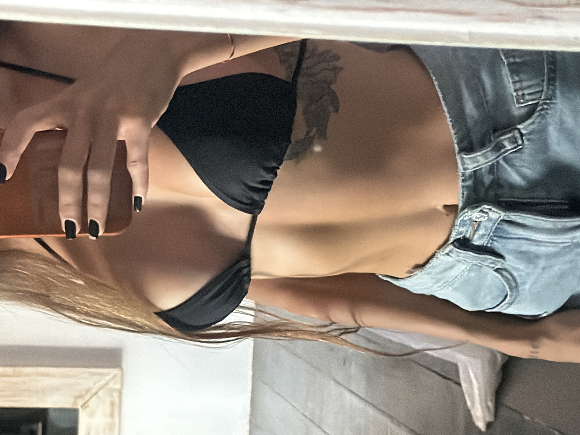 A woman with blonde hair and dark nail polish is taking a selfie in a mirror. She is wearing a black bikini top and light blue denim shorts. There is a tattoo on her chest. The location appears to be inside a room with a framed picture on the wall. The city and country are unknown, but potentially Unknown, Indonesia based on the request.