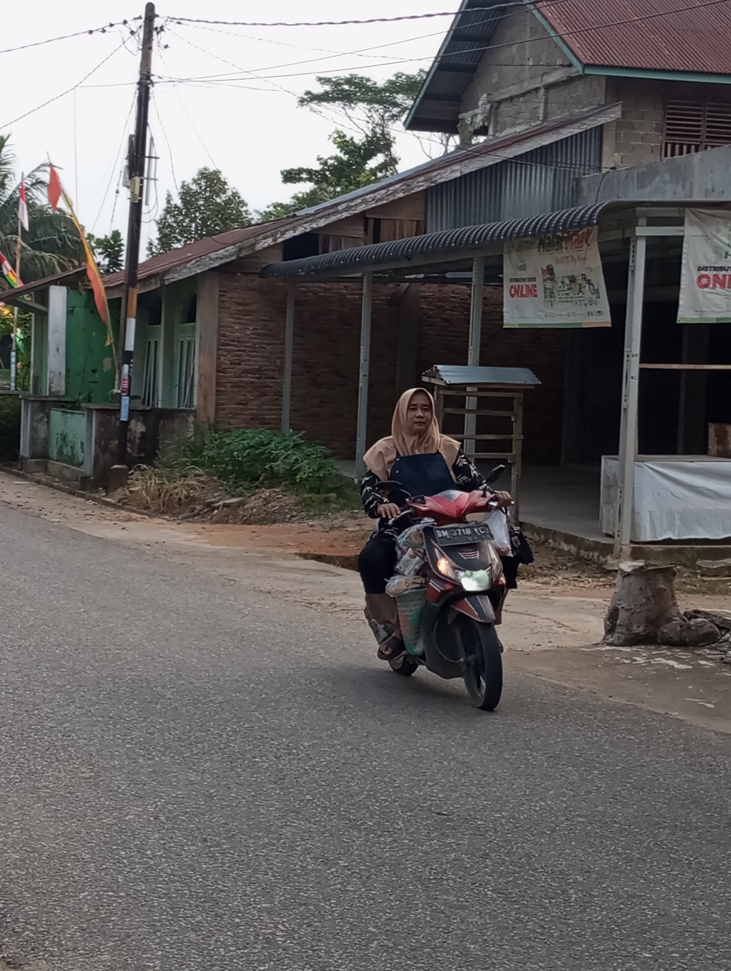 A woman wearing a headscarf rides a red and black scooter on a paved road in Pematang Reba, Indonesia. The woman has a dark blue apron over a patterned top and is wearing dark pants and sandals. The scooter has items in the front basket. She is looking forward. In the background, there are buildings with corrugated metal roofs and brick walls. Flags are visible on the left side of the image. The sky is overcast. The road appears to be in a rural or suburban area.