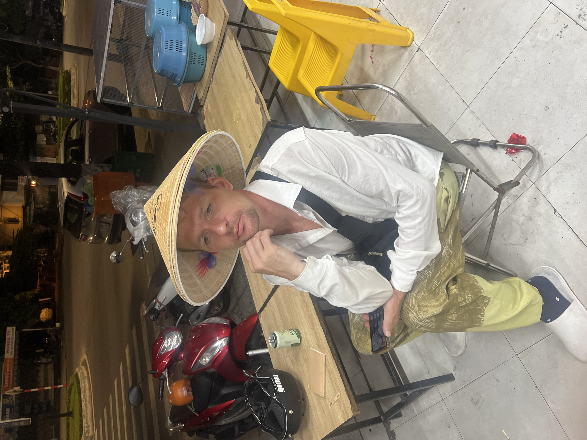 A man wearing a conical hat sits on a folding chair in front of a street food stall in Đà Nẵng, Vietnam. He is dressed in a white button-down shirt and patterned pants, holding a smartphone in his left hand. On the wooden table next to him are a motorcycle helmet, a can of beer, and a small notebook. Behind him, several red and white motorbikes are parked, along with yellow plastic chairs and blue plastic baskets. The scene appears to be at night, with streetlights illuminating the area.