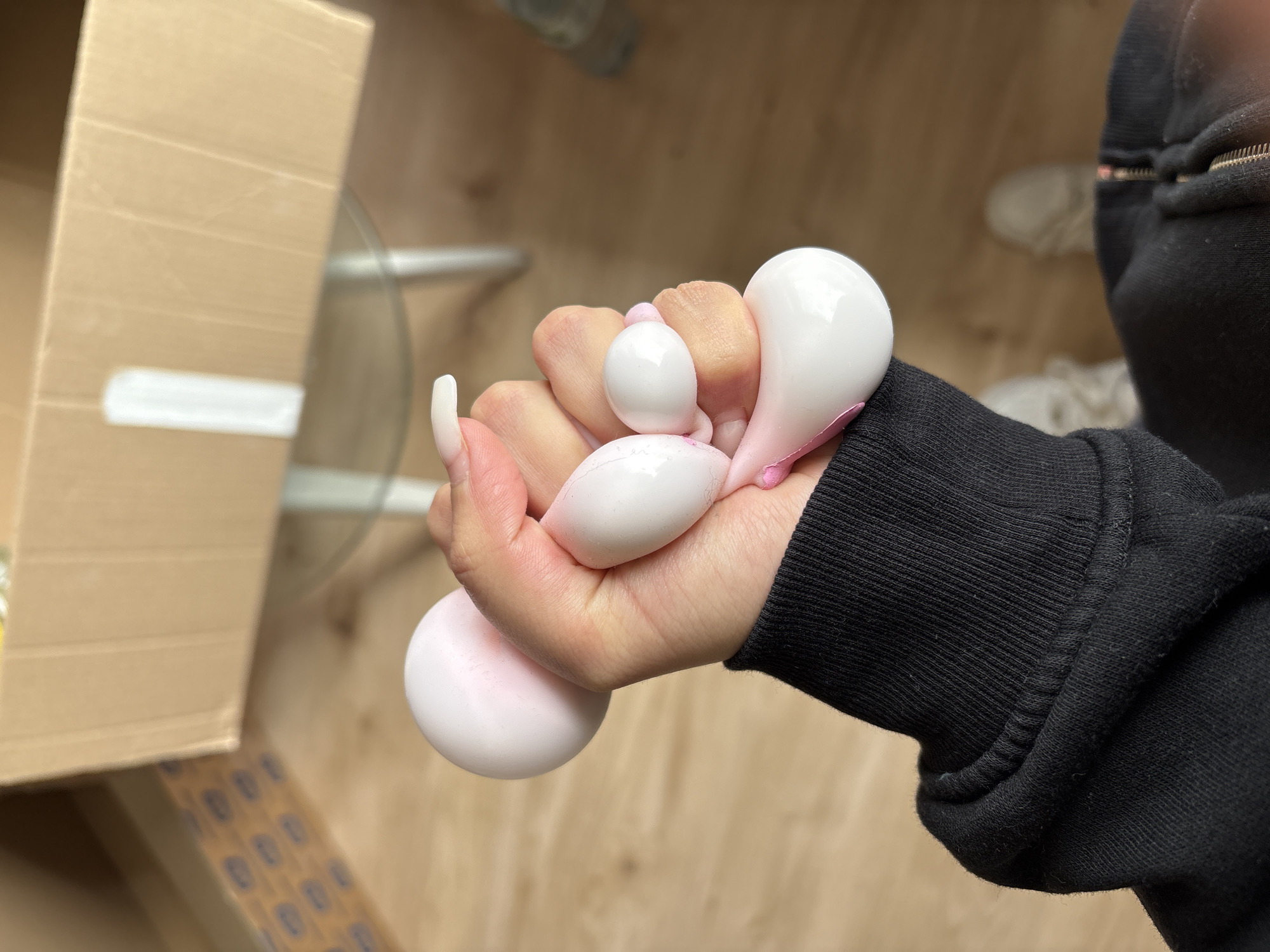 A human hand, with elongated fingernails, clutches several white stress balls with pink accents. The hand wears a black hoodie. The background includes a cardboard box and a table with a glass top. The location is Mönchengladbach, Germany.