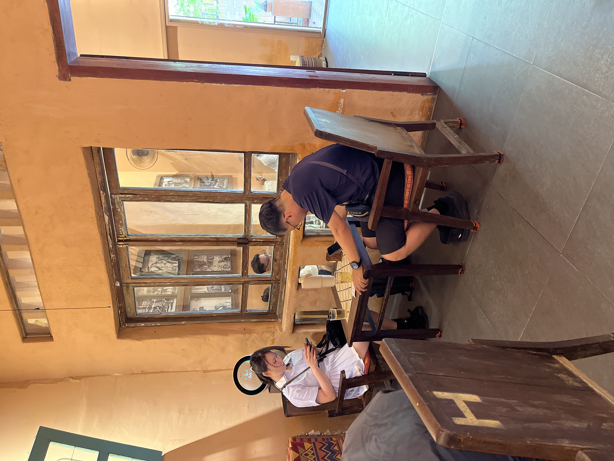 Captured in Đà Nẵng, Vietnam, the image presents an indoor scene, likely a cafe. Two individuals are seated at wooden tables with hinged leaves that can be folded up to make a work space. A man, wearing a dark blue t-shirt and shorts, is leaning forward at one of the tables. Seated across from him is a woman in a white dress, looking at her phone. A window is in the background. The cafe has a simple aesthetic, with walls painted in shades of brown.