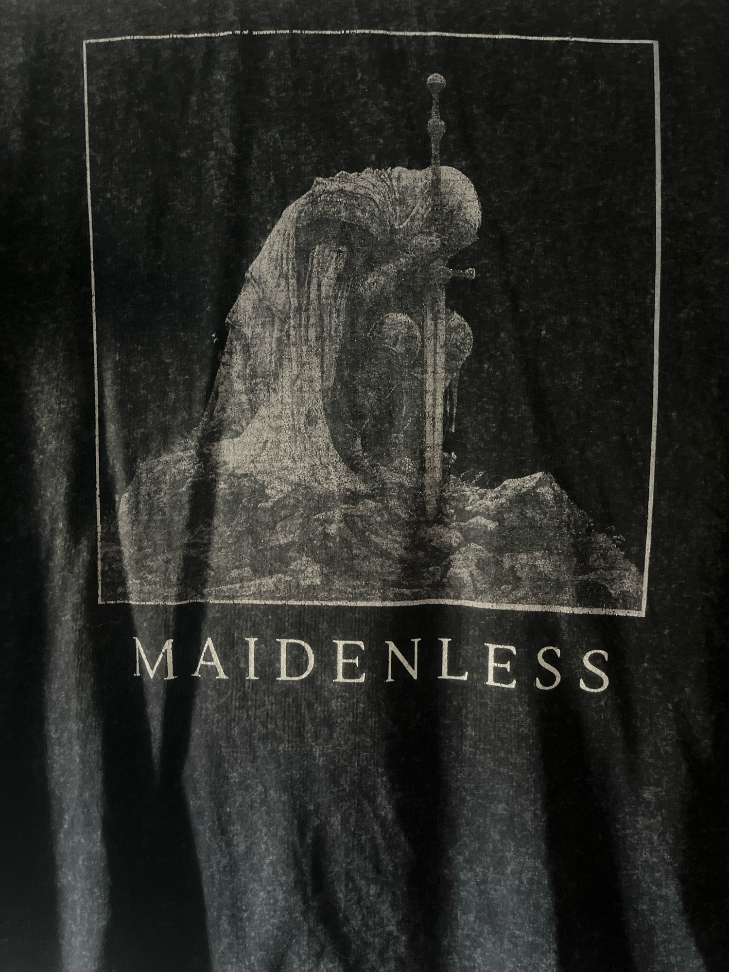 The image displays a black t-shirt with a distressed graphic print. The print features a monochrome, artistic rendering of a robed figure kneeling with a sword plunged into the ground before them. The figure appears to be facing away from the viewer, with their head bowed. The artwork is contained within a rectangular border. Below the graphic, the word "MAIDENLESS" is printed in white, serif capital letters. The fabric of the t-shirt has a textured, faded appearance, and there are shadows cast across the print, suggesting it is an indoor photograph. There is no indication of the city of Kristianstad, Sweden, or any specific location within it.