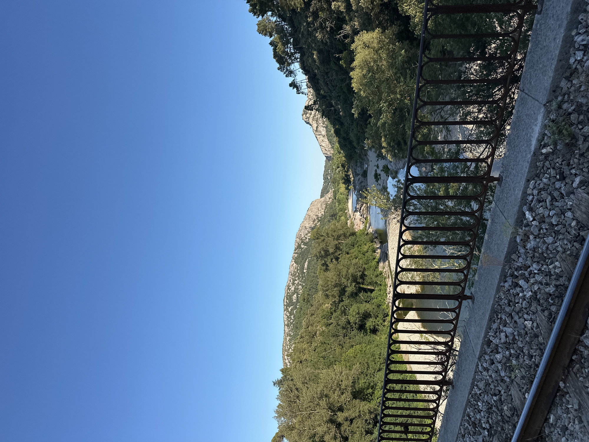 An elevated, wide-angle shot captures a tranquil river winding through a rocky, tree-lined valley under a clear blue sky. In the foreground, a rustic, dark metal railing stands on a concrete ledge, behind which are train tracks and ballast. The railing overlooks the river and the surrounding landscape of lush green trees and rugged, rocky hills that recede into the distance. The river is shallow, with visible rocks and sandbars. The scene is bathed in the warm light of late afternoon, casting long shadows. This appears to be the Gardon River as it flows through the Cévennes region near Anduze, France, possibly from a vantage point near the town's old railway station. There are no people or animals visible in the image.