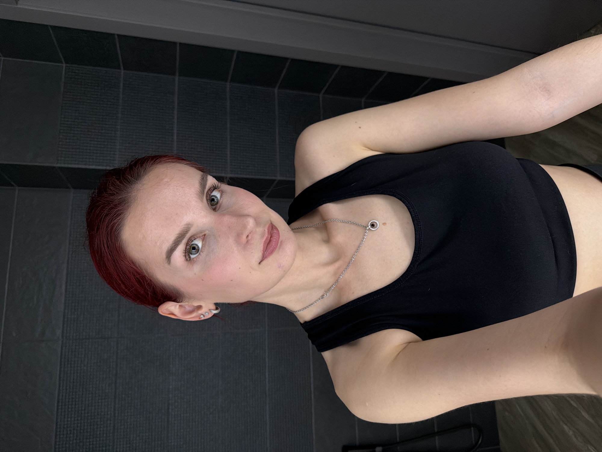 A woman with short, red hair is positioned in front of a dark gray tiled wall. She is wearing a black tank top and a silver necklace with a circular pendant. Her gaze is directed at the camera. The setting appears to be indoors, possibly a bathroom or tiled space. The location, Chaikovsky, Russia, does not impact the description.