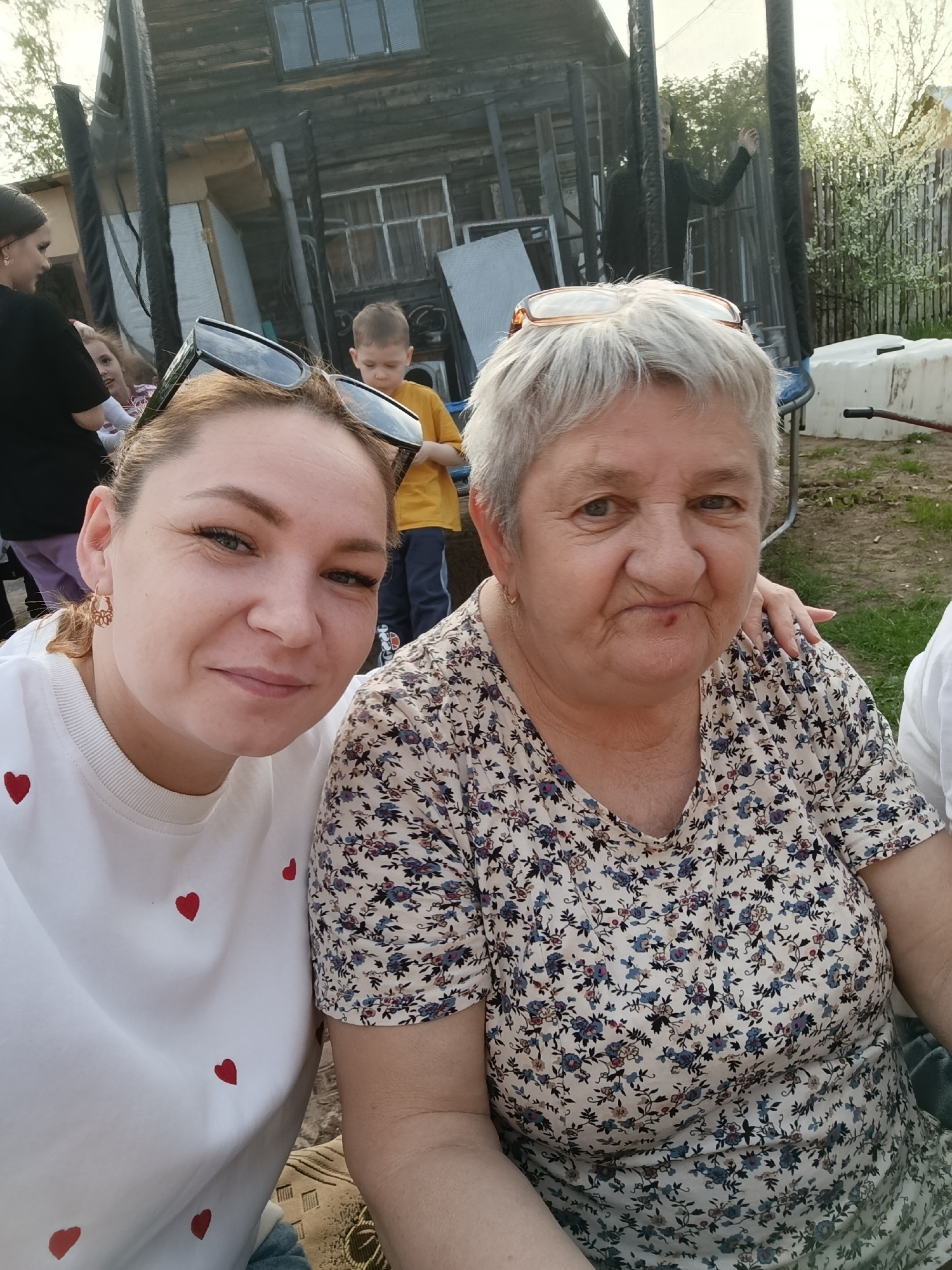 The image captures a selfie of two women, likely a mother and daughter, taken outdoors in what appears to be a backyard setting. The younger woman, on the left, has light brown hair pulled back, wearing a white t-shirt with small red hearts. She is smiling slightly. The older woman, on the right, has short grey hair and is wearing a floral print top. She also has a slight smile. Behind them, a young boy in a yellow shirt is standing near a trampoline and a partially visible wooden structure resembling a shed or outbuilding. Other indistinct figures can be seen in the background, along with a white container and hints of greenery and fencing. The location is specified as the city of Chaikovsky, Russia.