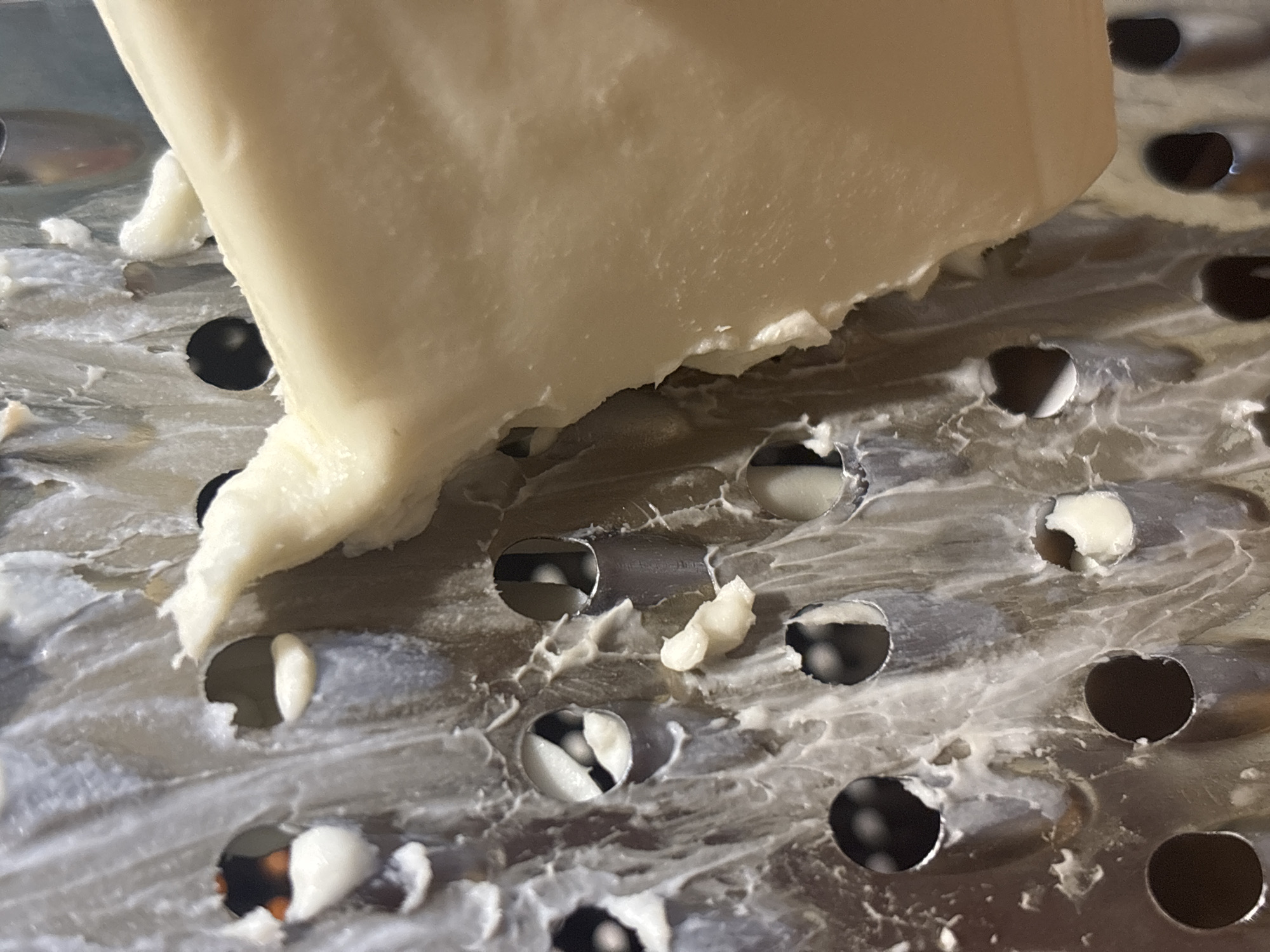 A close-up, high-angle shot shows a piece of pale yellow cheese being grated on a metal grater. The grater has multiple holes of varying sizes, and shredded cheese is visible clinging to its surface. The background is blurred, but appears to be a domestic kitchen setting. There is no indication of the location in Kremenchuk, Ukraine.