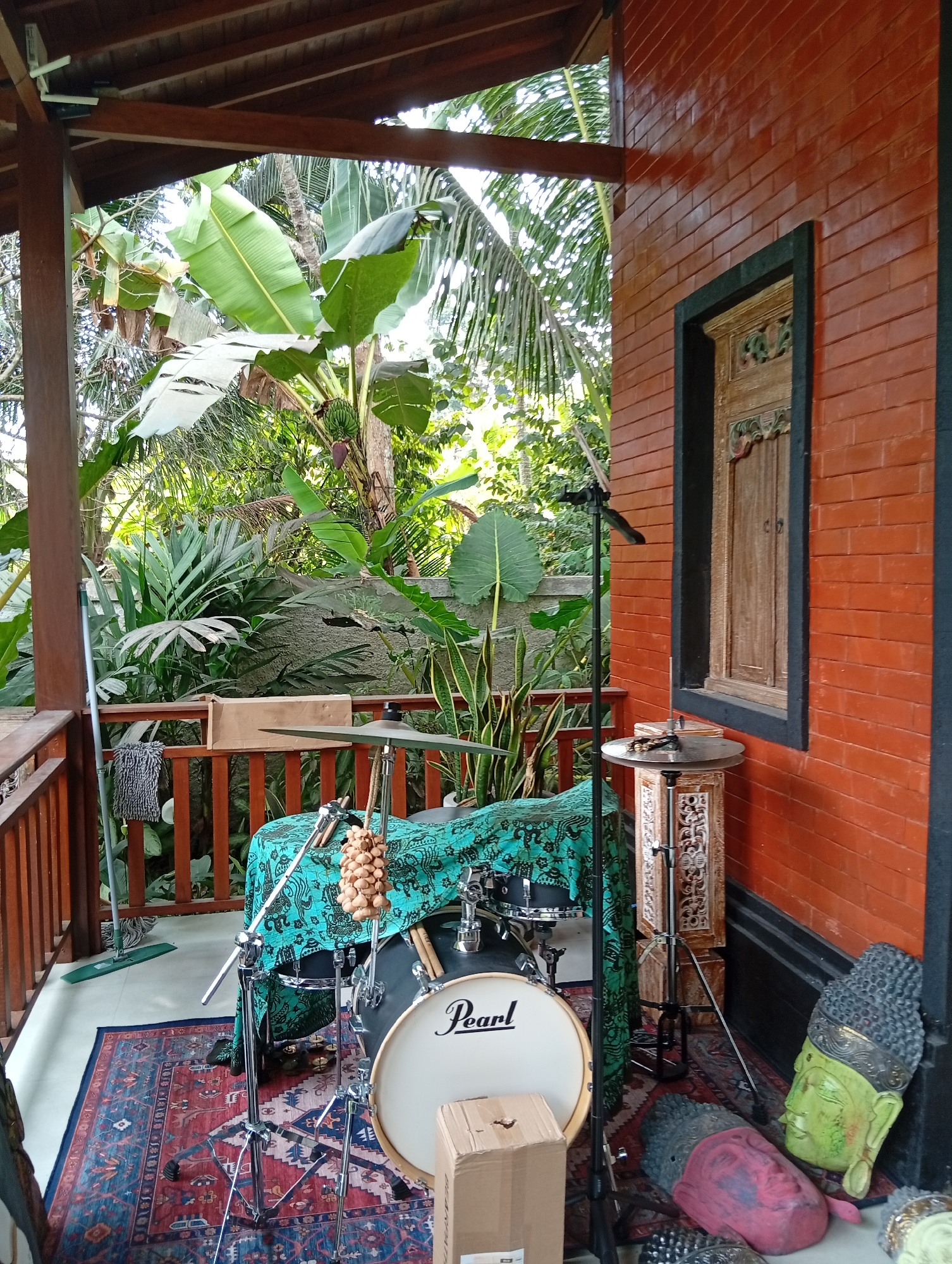An outdoor covered patio in Ubud, Indonesia, features a drum set covered with a green patterned cloth. To the right of the drum set, a brick wall with a carved wooden window is visible. The patio is decorated with two Buddha head statues, one green and one red, lying on the floor. A rug with a red, blue, and white pattern is placed under the drum set. Lush tropical foliage, including large green leaves and palm fronds, surrounds the patio. A mop and a cardboard box are also present.