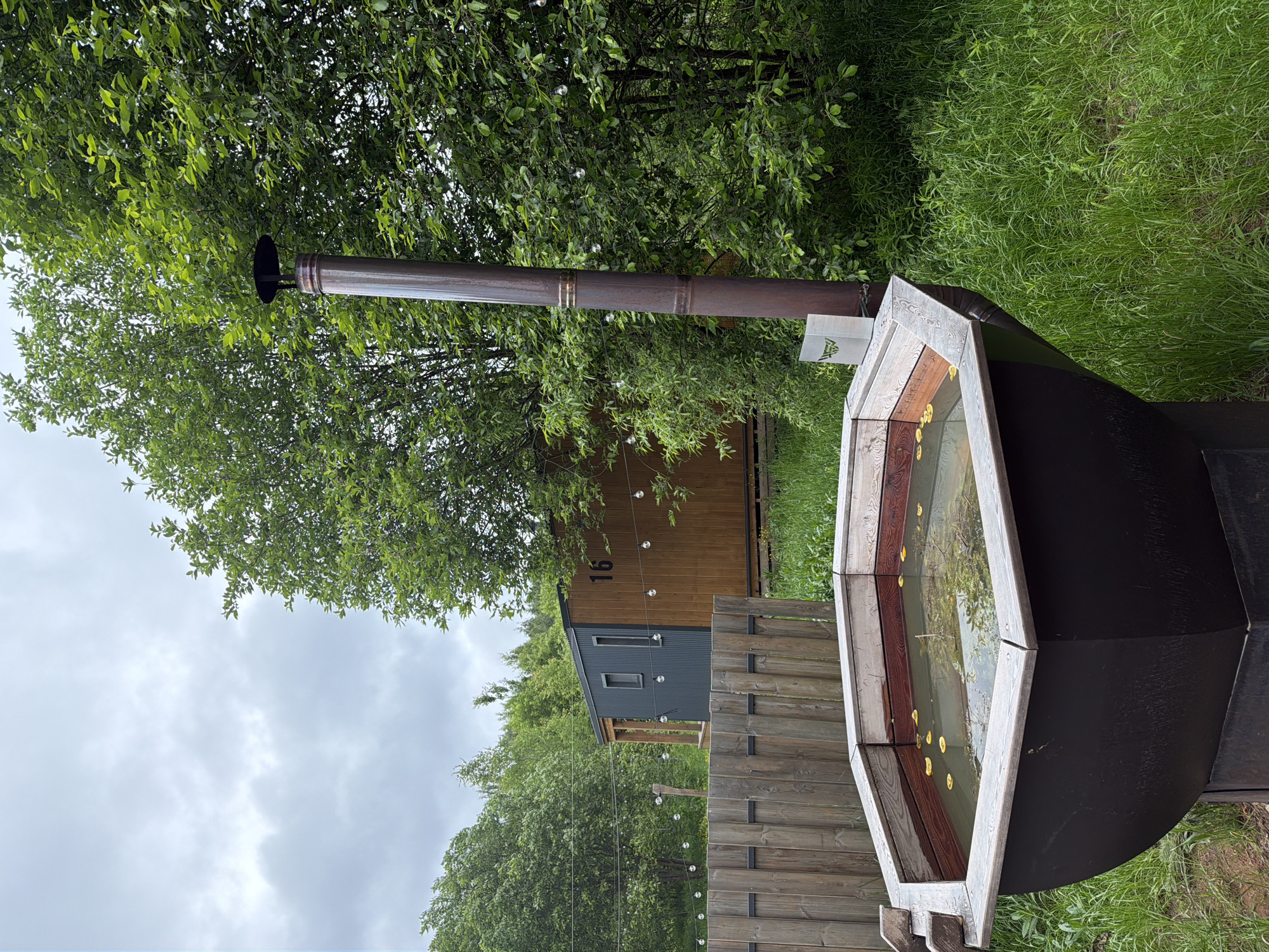 The image captures an outdoor, rustic scene in Vozhoiskii, Russia, under an overcast sky. In the foreground, a dark-colored outdoor hot tub with a light-stained wooden rim is prominent. The water within the tub contains several floating yellow items, resembling petals or small fruits. A wooden fence, constructed from horizontal planks, runs along the left side of the frame, behind the hot tub.
Rising behind the hot tub is a tall, dark metal chimney pipe. A small, white rectangular sign with a green, triangular logo (resembling mountains or an initial "A") is hanging from the chimney. Further in the midground, a small cabin or dwelling is visible. It features a lower section clad in dark corrugated metal and an upper section made of natural wooden planks. The number "16" is clearly marked on the wooden facade of the cabin. String lights with exposed bulbs are strung across the scene, extending from the left towards the cabin.
The background consists of dense, lush green trees and tall green grass, indicating a natural, possibly wooded, environment. The sky is cloudy and grey, suggesting an overcast day. No people are present, and no specific activity is occurring, but the setup implies a setting for relaxation and leisure.