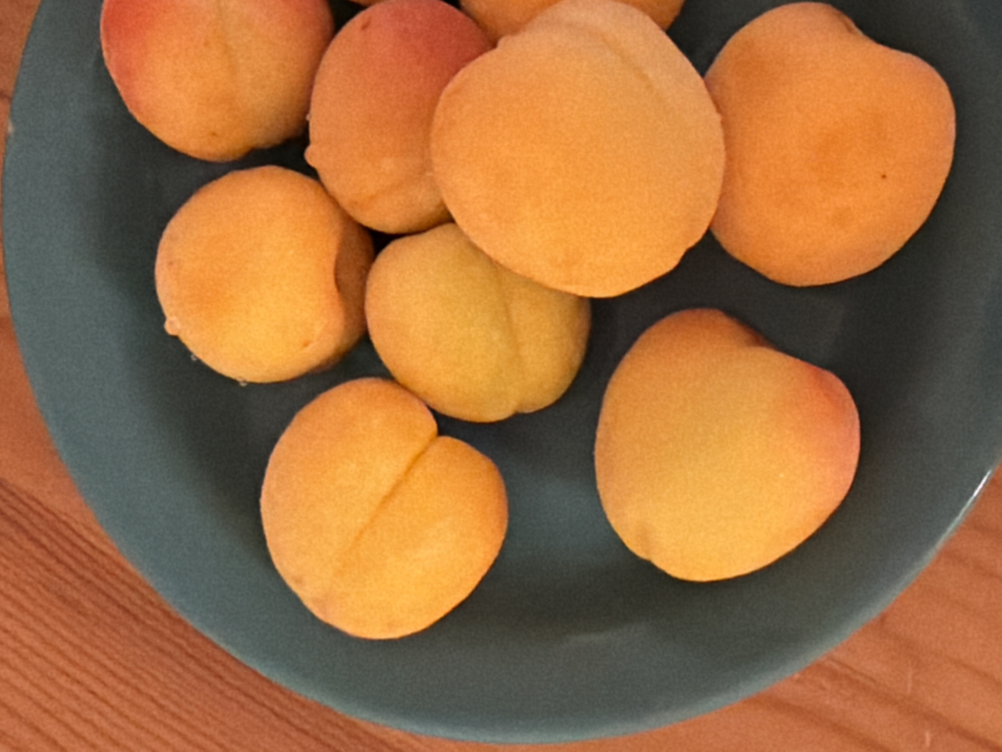A close-up, high-angle shot displays a collection of apricots on a dark teal plate, which rests on a light brown, wooden surface. The apricots vary slightly in color, ranging from yellow to orange, and are arranged haphazardly on the plate. The scene is suggestive of food.